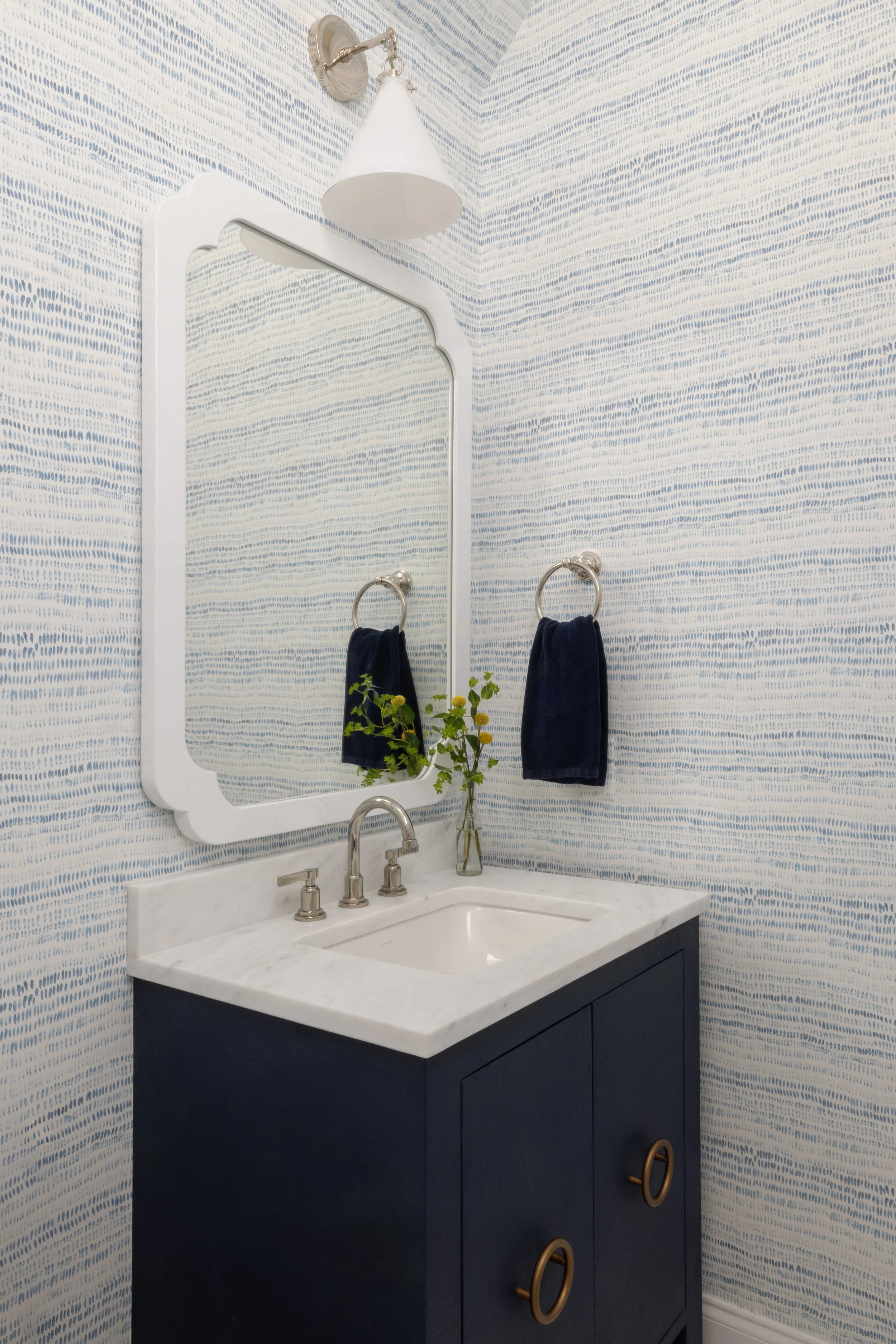 Small bathroom with patterned blue and white wallpaper, a dark blue vanity with a white marble top, a rectangular mirror, a glass vase with green foliage, navy blue hand towels on silver rings, and a white wall-mounted lampshade.
