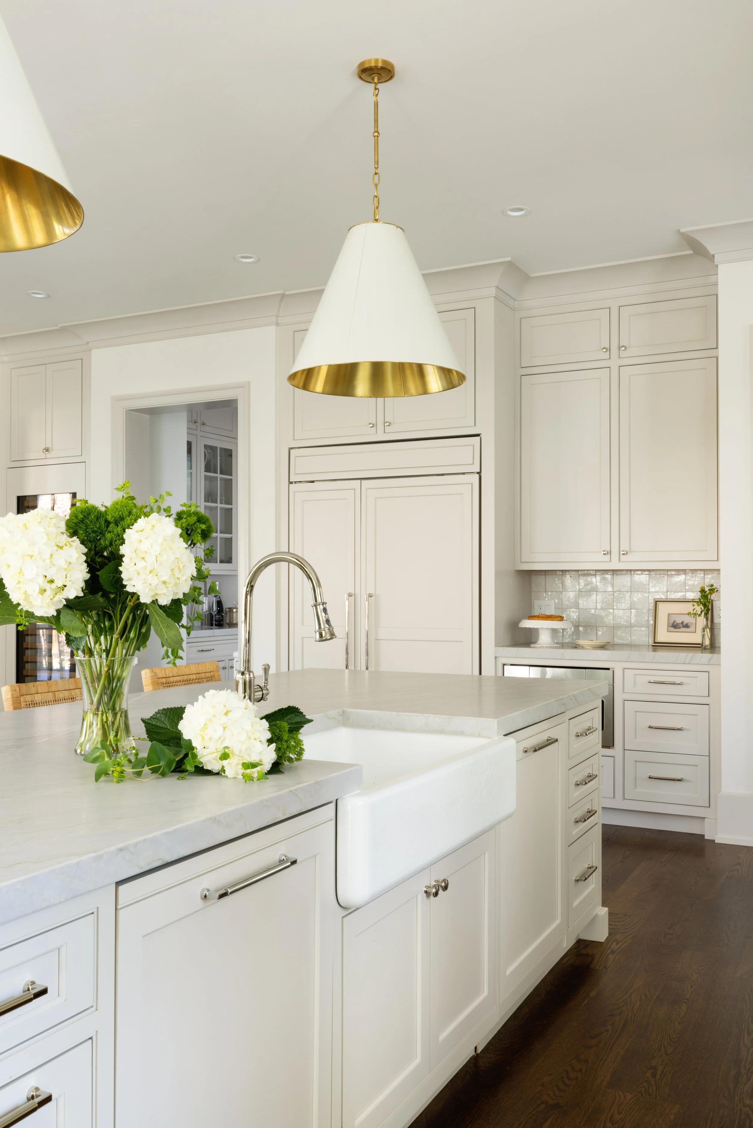 A bright, modern kitchen with white cabinets, a marble countertop, a white farmhouse sink, and a vase of white hydrangeas on the island. There are gold accents on the hanging light and cabinet hardware, with dark wood flooring.