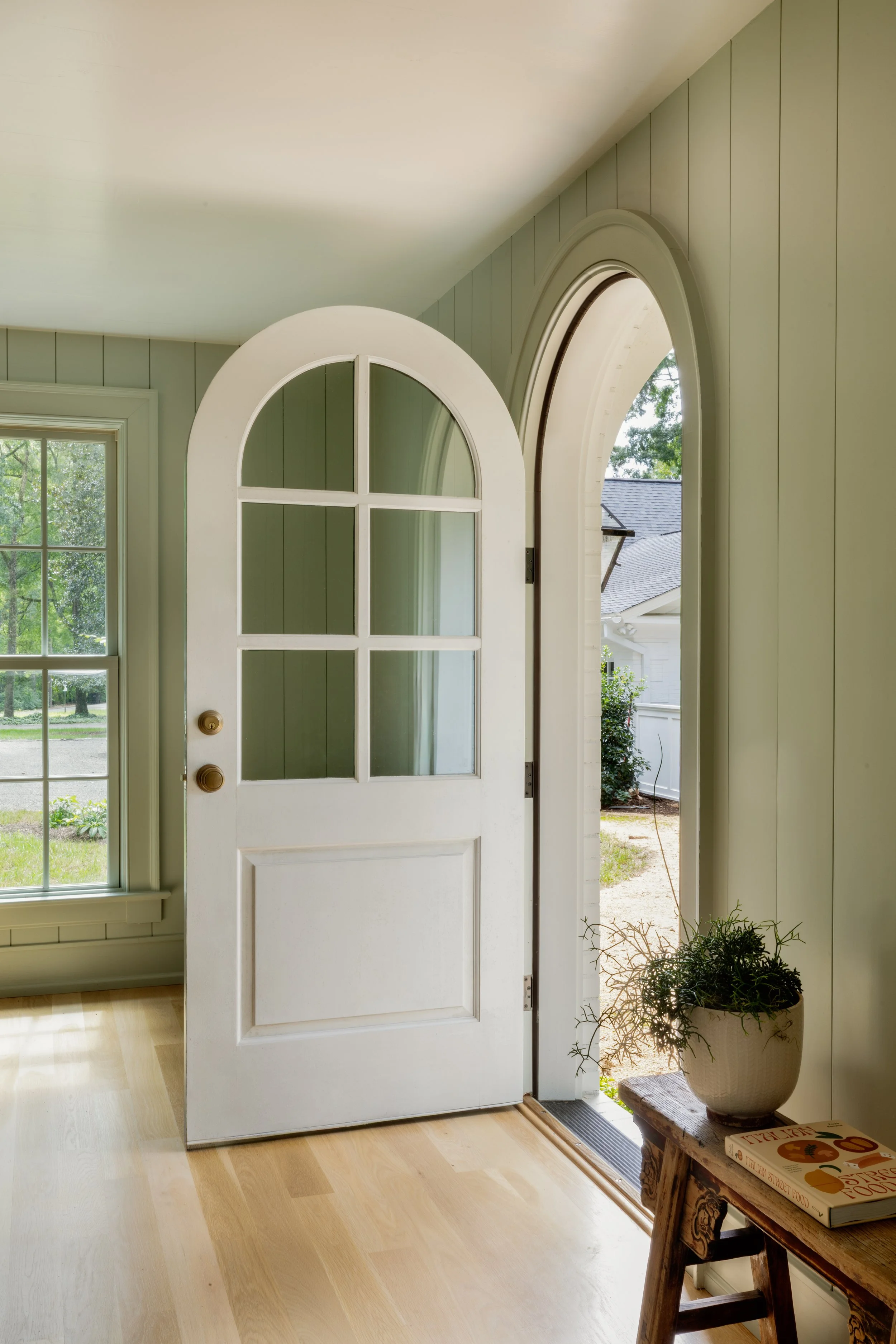 Open white door with an arched window leading outside, in a sunlight-filled room with wood flooring, a window on the left, and a small table with a potted plant and a book on the right.