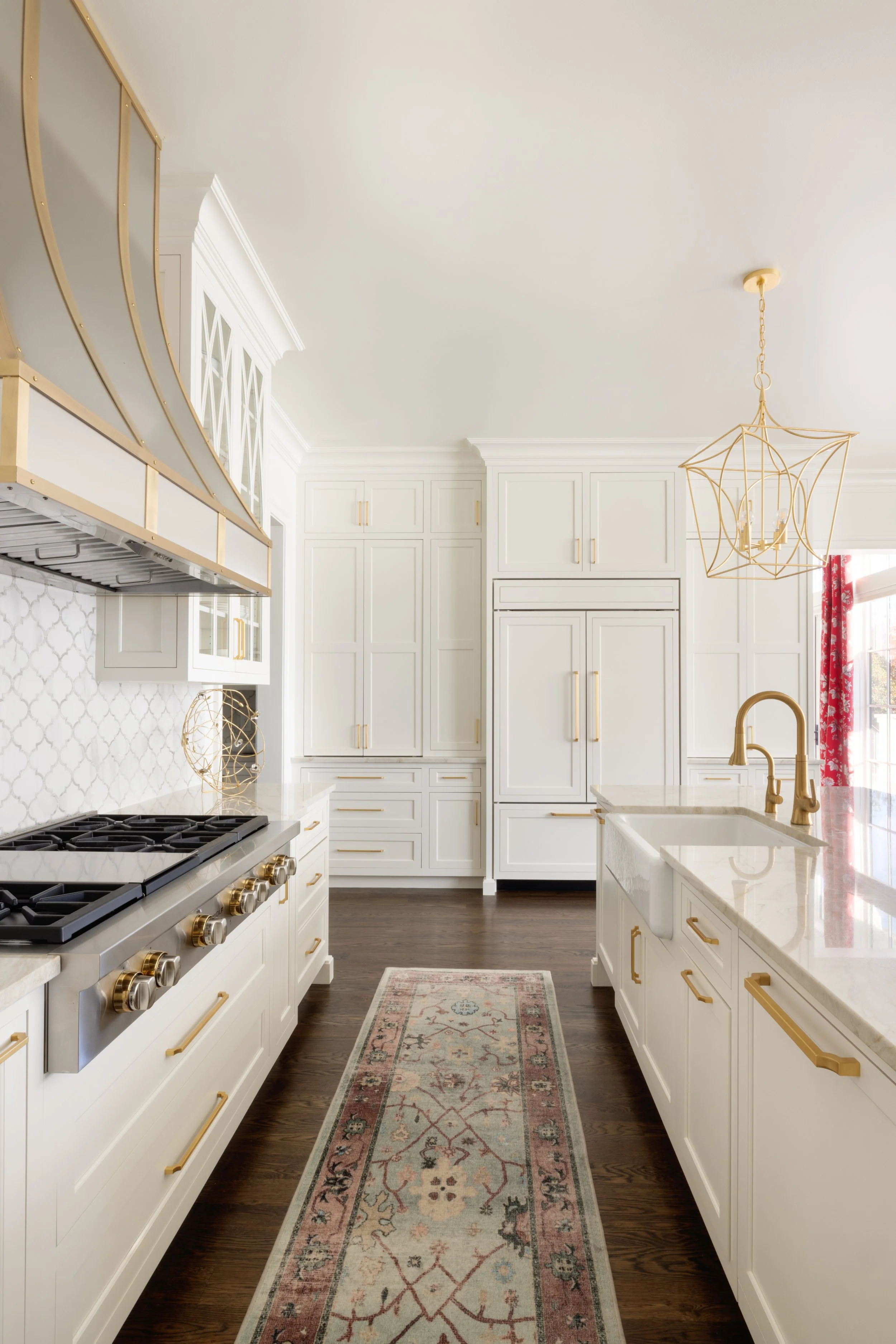 Bright modern white kitchen with gold accents, a central island with a white marble countertop, a gold-rimmed rug on dark hardwood floor, and a geometric gold pendant light hanging from the ceiling.