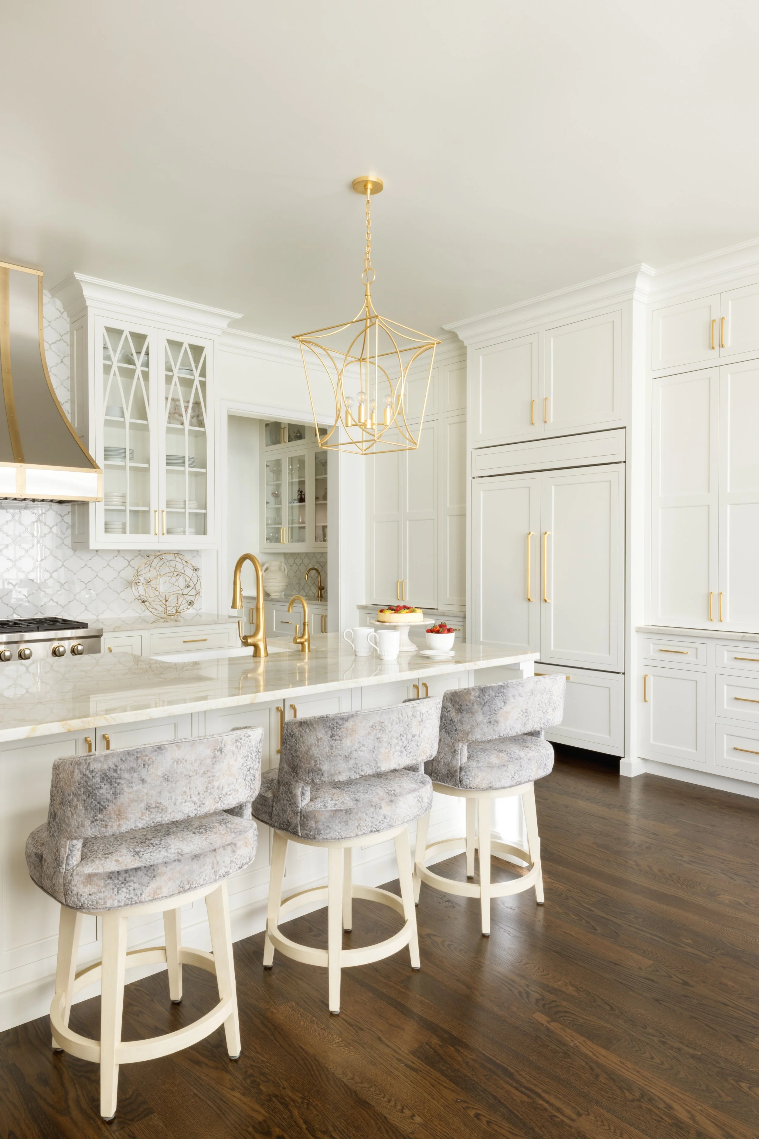 Modern white kitchen with gold accents, three bar stools with gray velvet upholstery, brown hardwood floors, and a geometric gold chandelier.