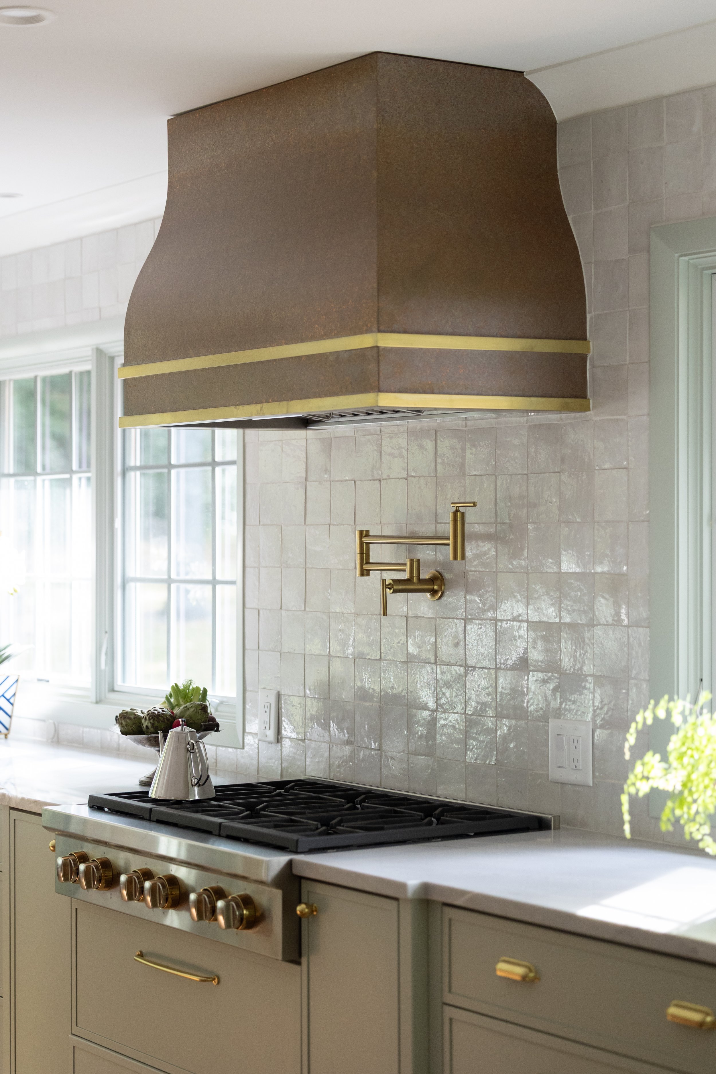 Kitchen with beige cabinets, a gas stove, a brass wall-mounted faucet, a tiled backsplash, and a large metallic range hood above the stove.