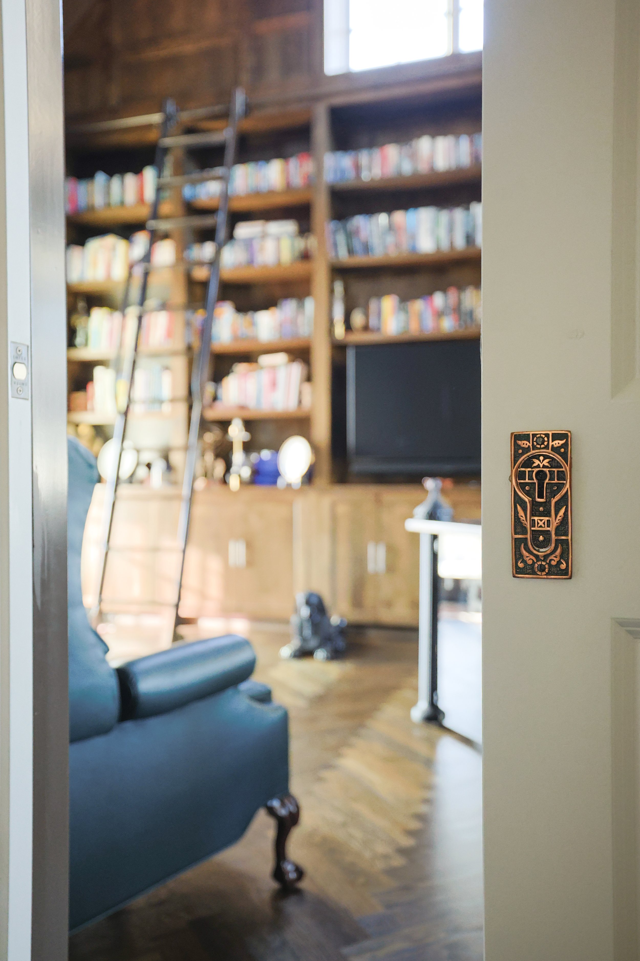 Interior view of a cozy living room or library with bookshelves filled with books, a ladder on the shelves, a leather armchair, and a wooden floor.
