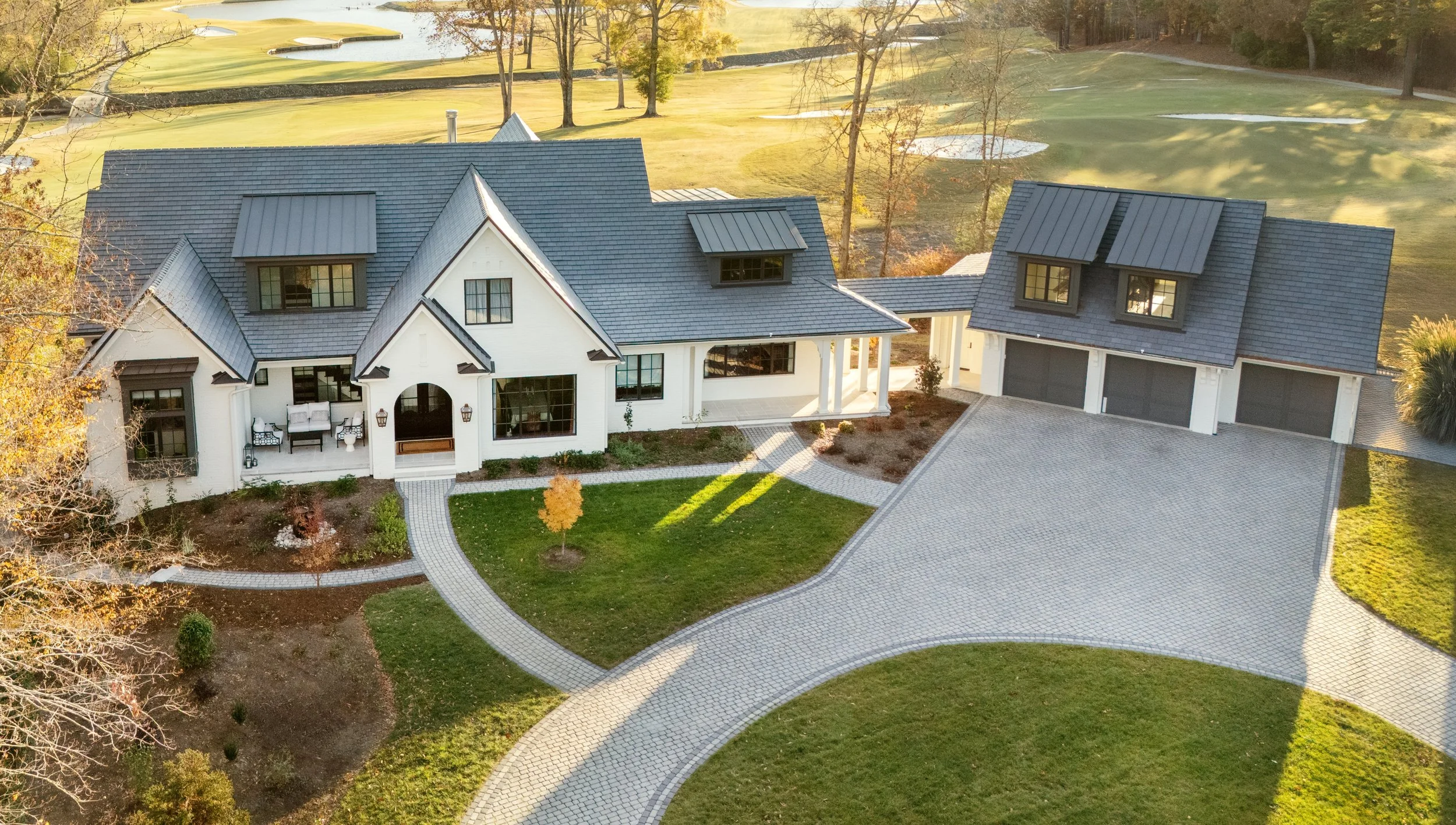 A large white house with black accents and a blue roof, featuring a covered porch with outdoor seating, surrounded by a landscaped yard with a curved paved driveway and a golf course in the background.