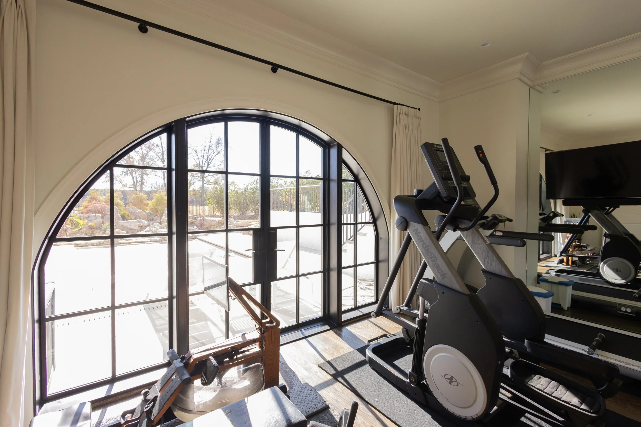 Home gym with treadmill, elliptical, and outdoor view through large arched window, with outdoor landscape including trees and rocks