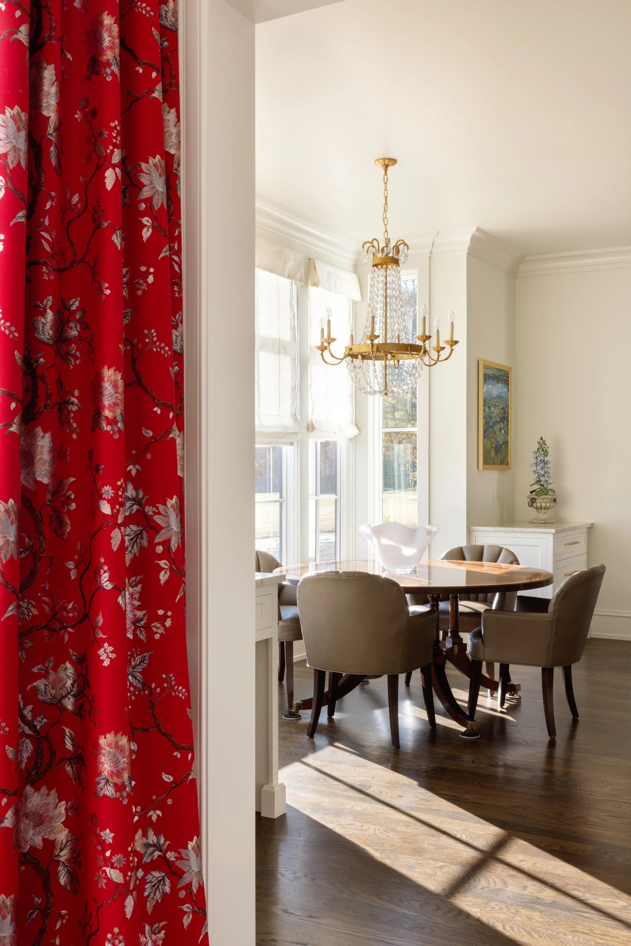 A dining room with sunlight streaming through large windows, a chandelier hanging above a round wooden table, surrounded by six upholstered chairs, a white buffet with decorative items, and a red floral curtain partially visible on the left side.