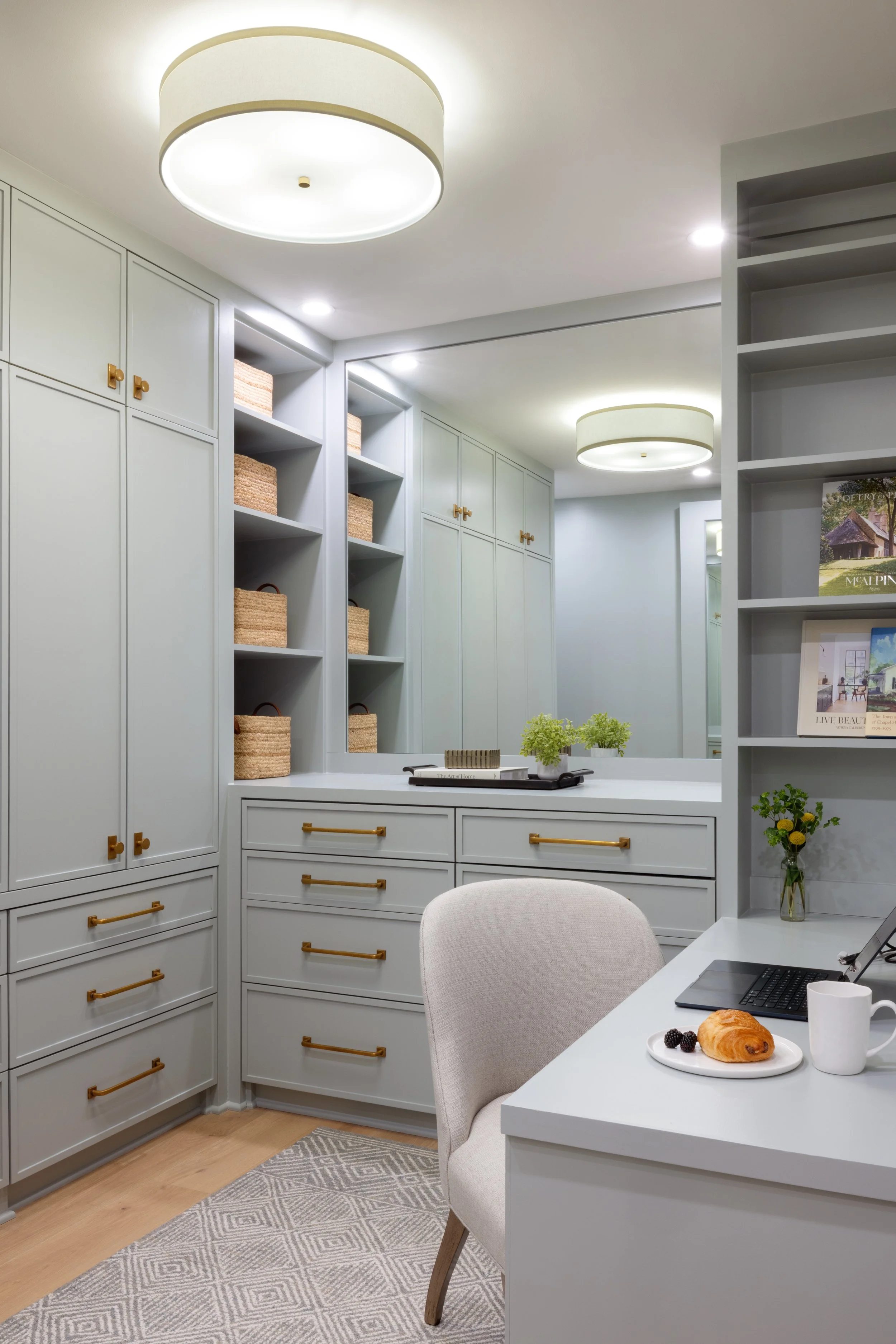 A cozy home office with white cabinetry, a large mirror, a desk with a laptop, a croissant, and a cup, and ceiling lights.