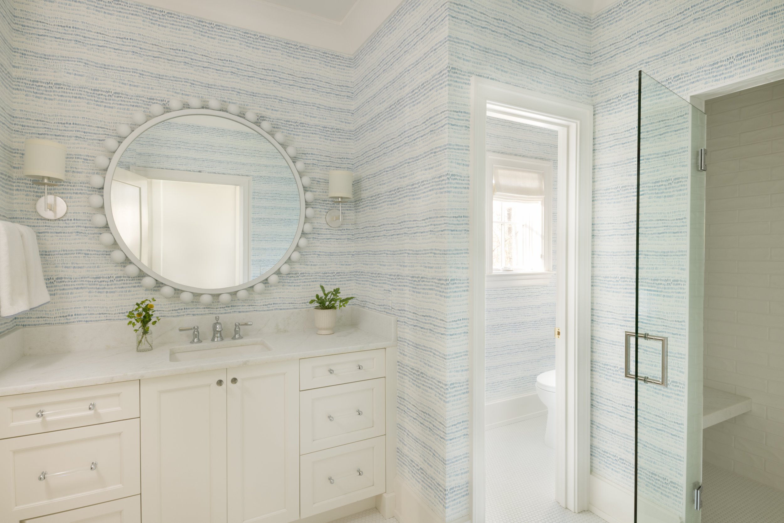 Bright bathroom with a white vanity with drawers and brass handles, a marble countertop, and a round mirror with a light bulb frame. There are two wall sconces with white shades on either side of the mirror. A small potted plant and a vase with flowe