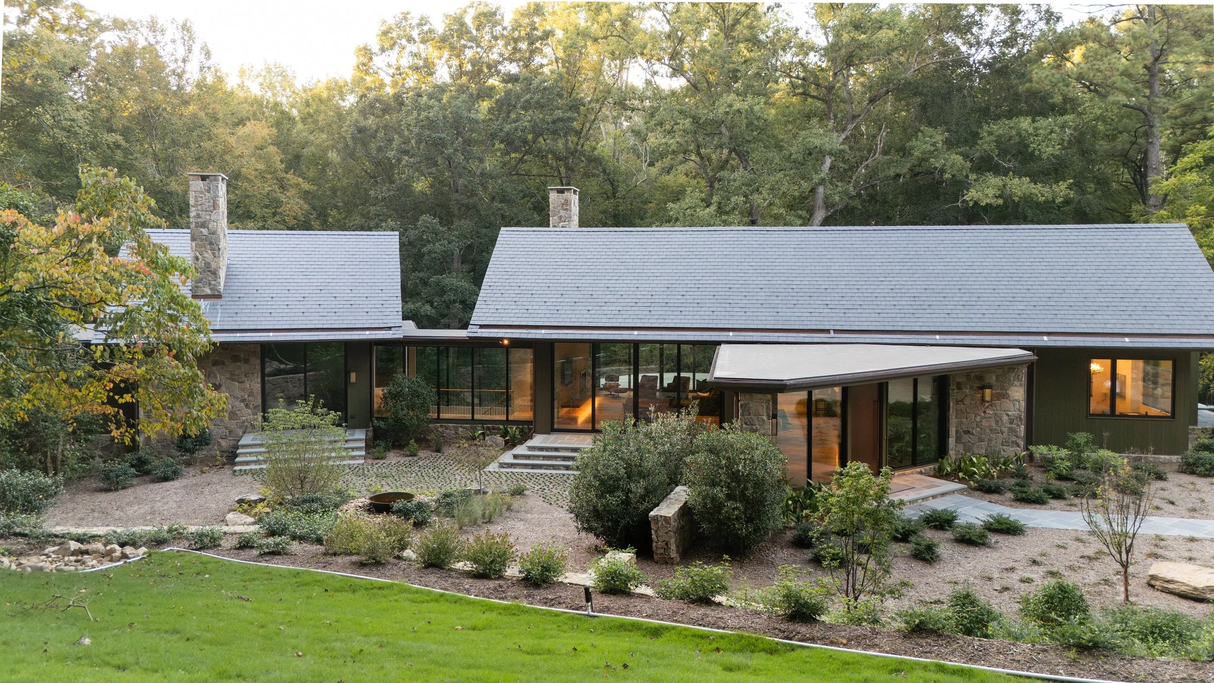 Modern house with stone and dark siding exterior, large windows, surrounded by landscaped garden with trees, bushes, and a well-maintained lawn, located in a wooded area.