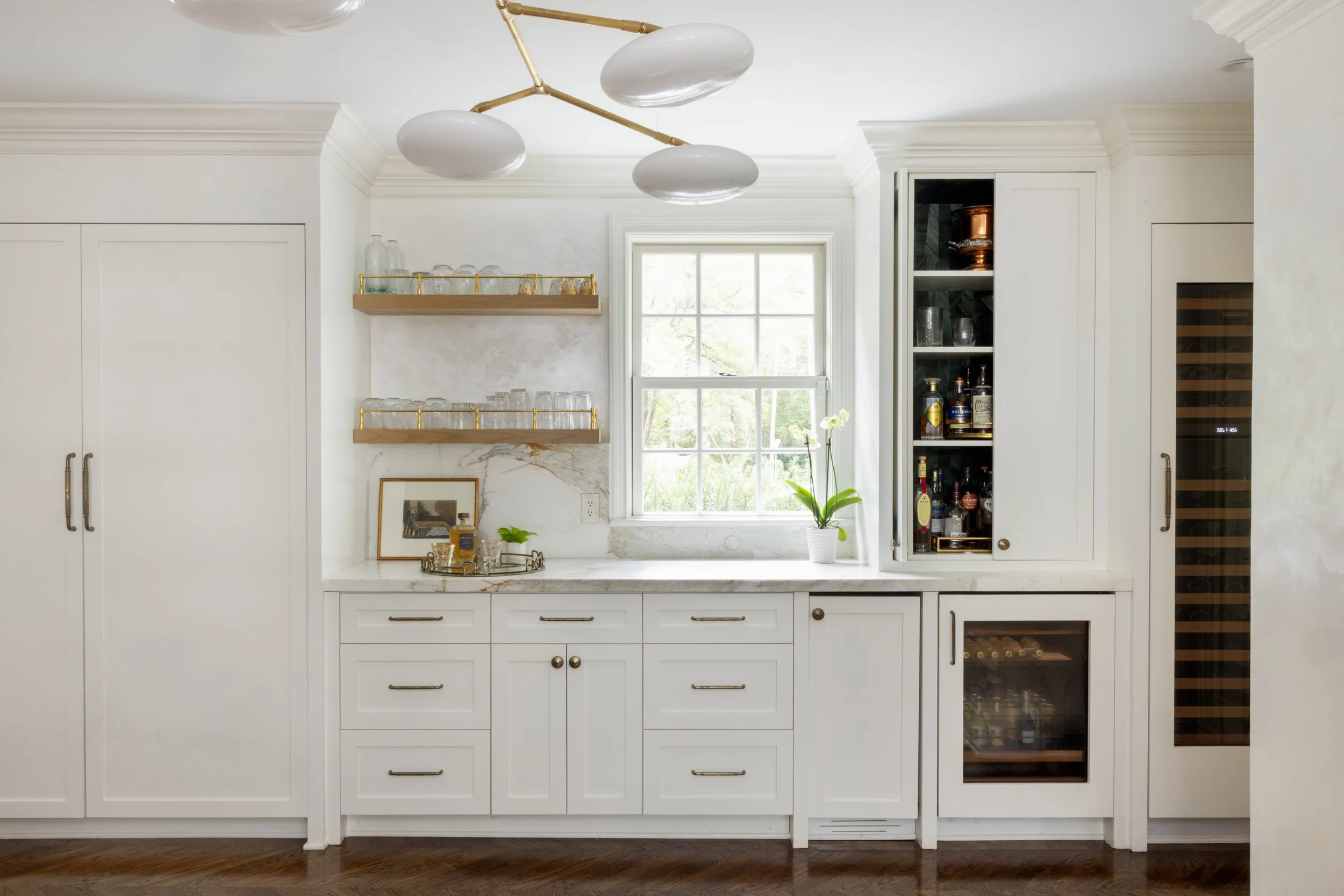 A bright modern kitchen with white cabinets, a marble countertop, open shelving with glassware, a window with a view of greenery, a small potted plant, and built-in wine and liquor coolers.