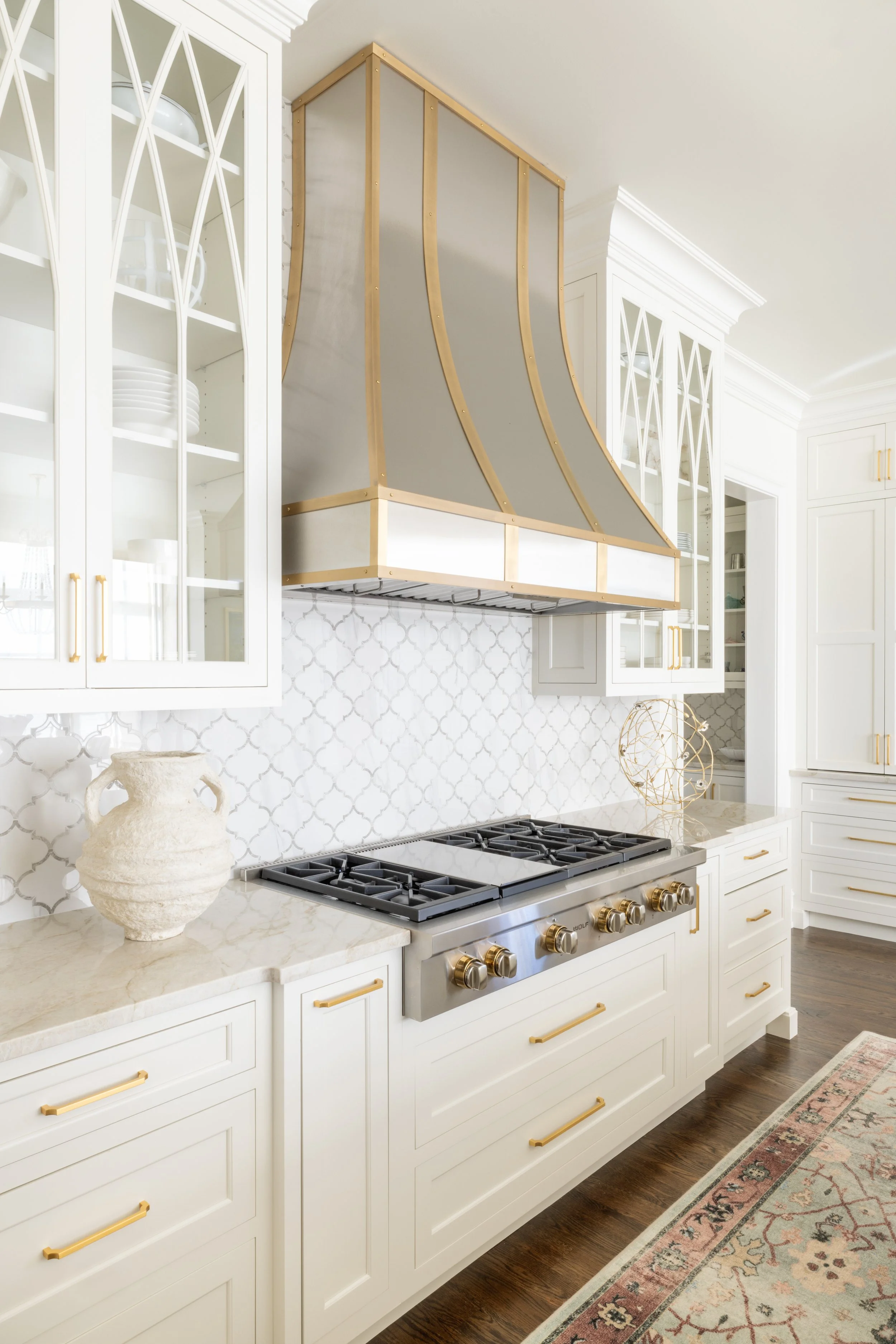 Kitchen with white cabinets, gold hardware, a marble countertop, and a decorative rug. There is a large range hood with a geometric design and a metallic finish over a stainless steel stove with gold knobs. White patterned backsplash and display cabi