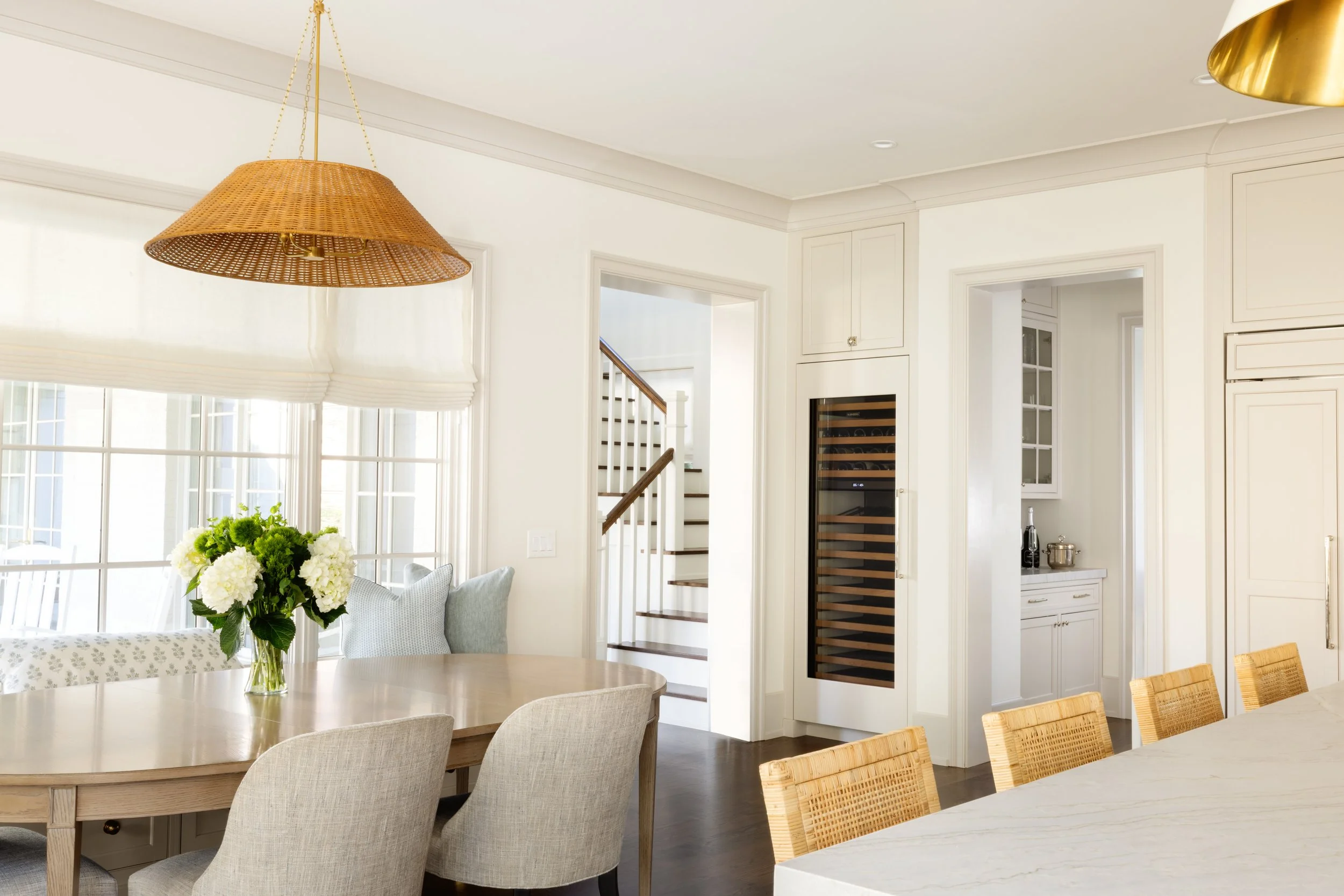 Bright dining room with a round wooden table, beige upholstered chairs, and a vase of white flowers. Large windows with white blinds, a staircase with white risers and dark treads, and a built-in wine cooler in the wall.