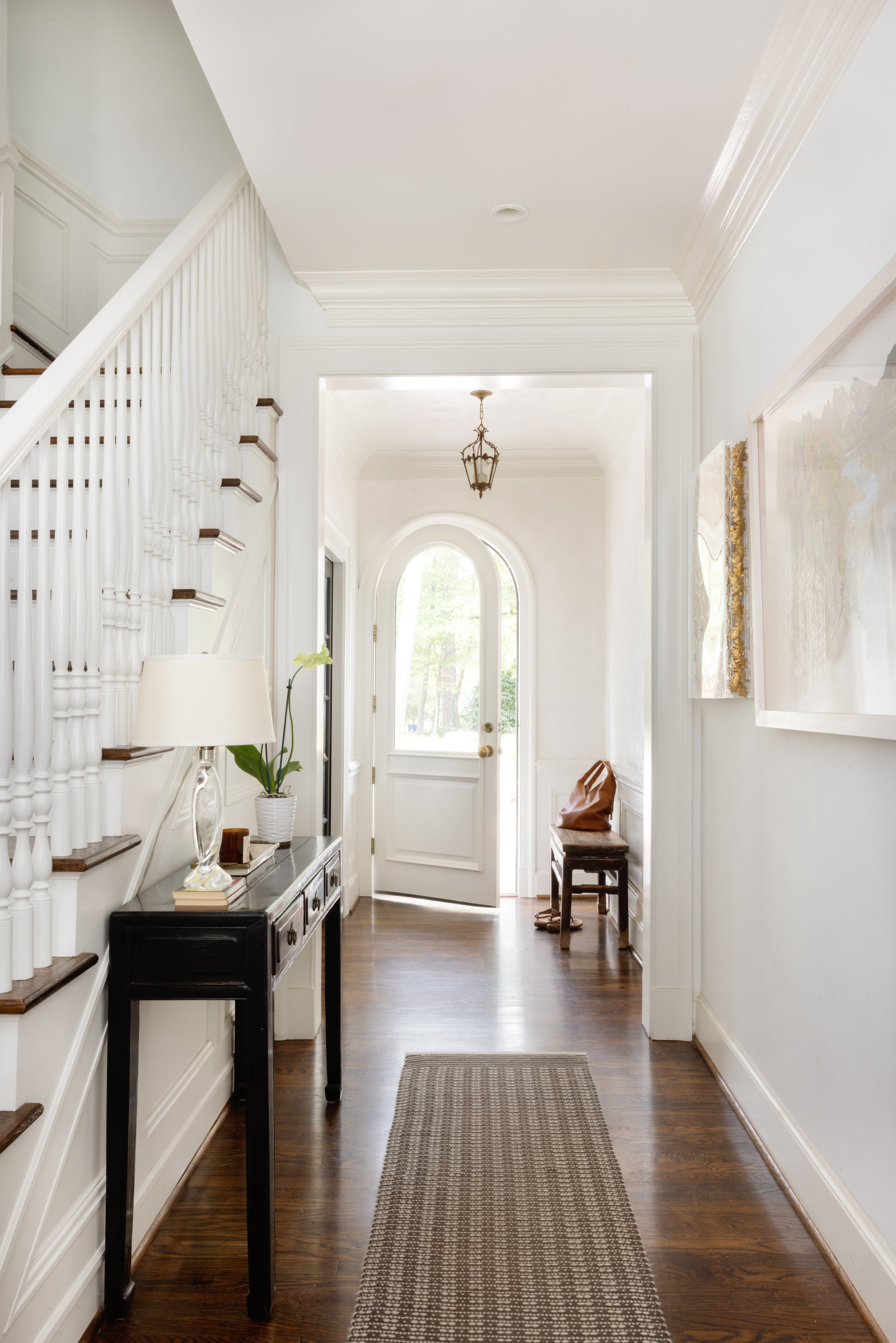 Bright entryway with a white front door, dark wooden floors, a black console table with a lamp and potted plant, and artwork on the wall.
