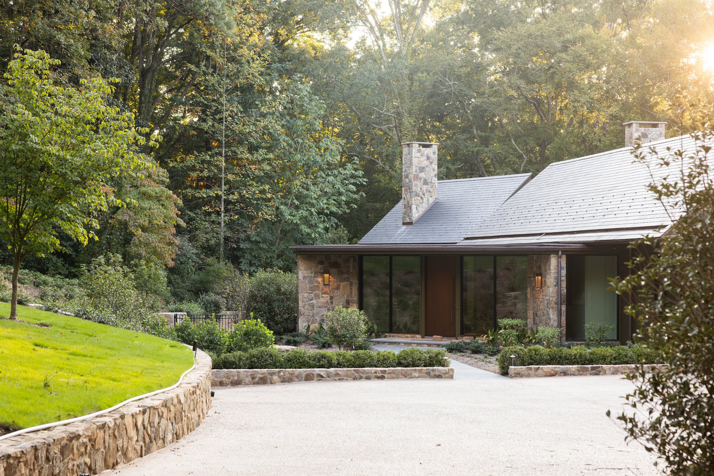 A modern house with stone and glass exterior, surrounded by greenery and trees, with sunlight shining through the trees.