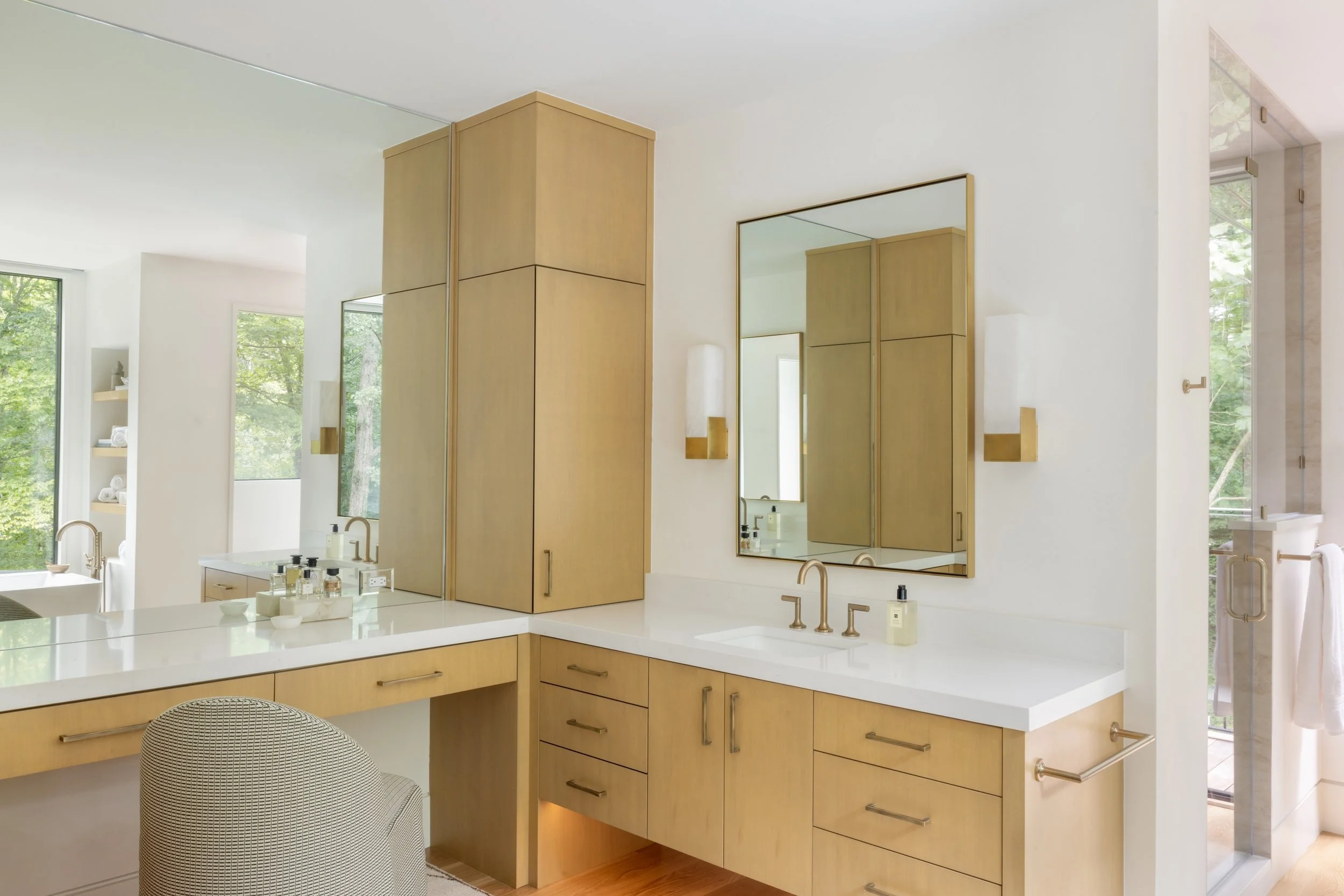 Modern bathroom with light wood cabinetry, white countertops, two gold faucets, and large mirrors. Natural light from windows shows greenery outside.