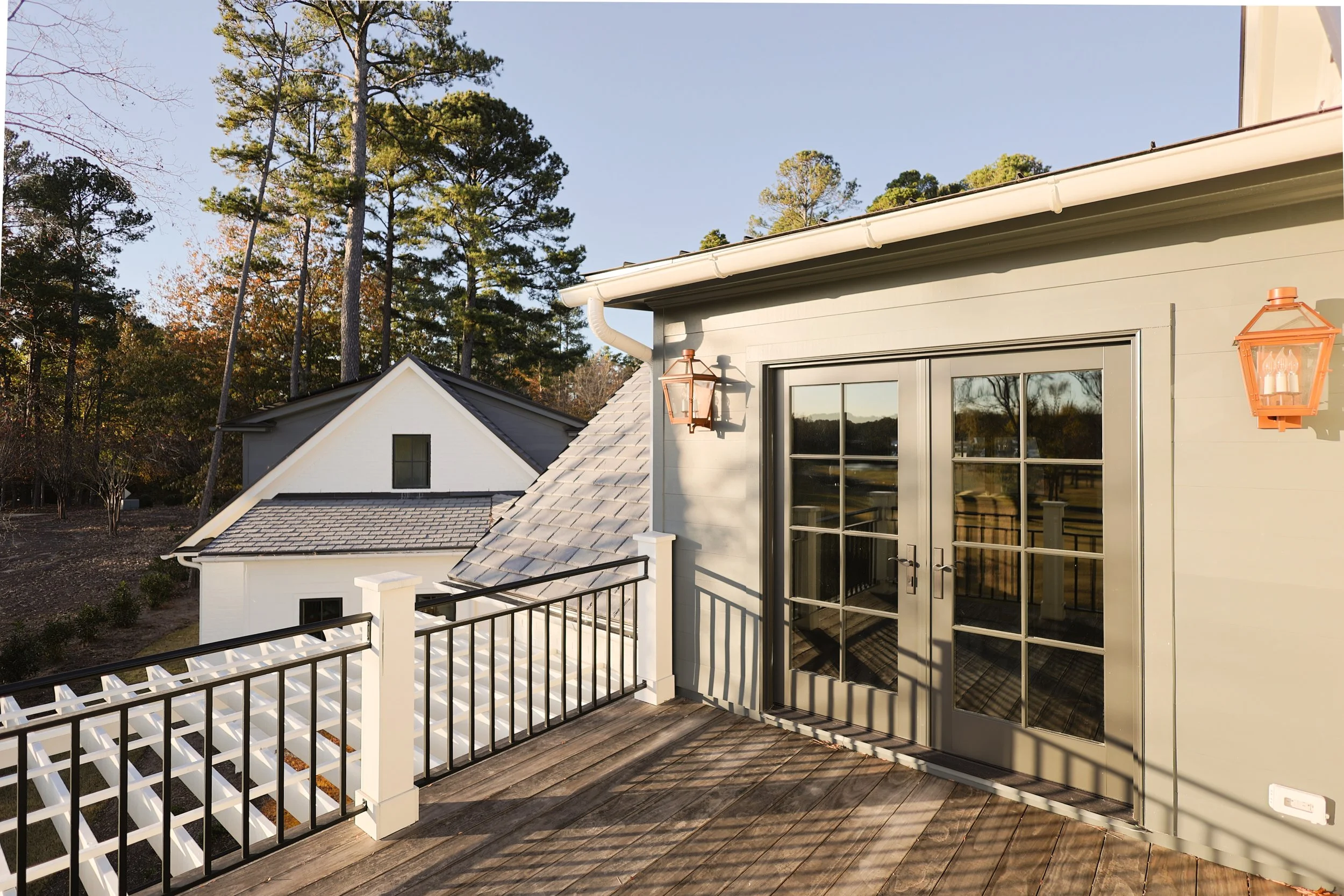 Exterior view of a house deck with black and white railing, French doors, and two wall-mounted lanterns. Trees and neighboring houses are visible beyond.
