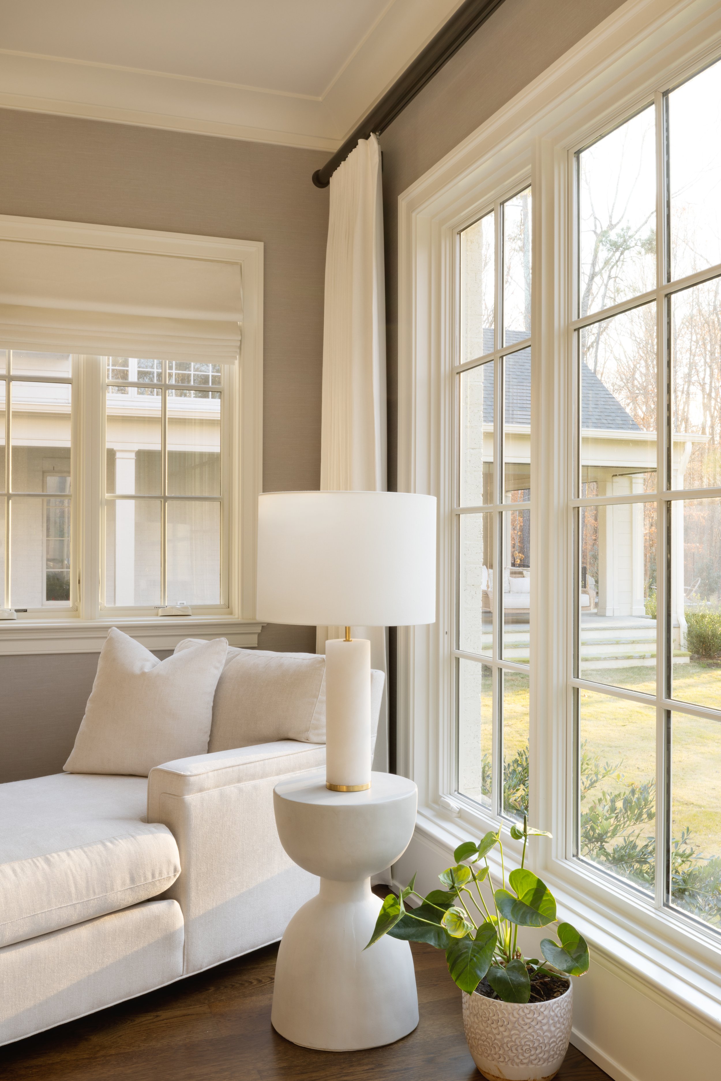 A bright living room corner with a beige sofa, a white table lamp on an abstract white side table, and a potted plant near large windows showing a lawn and neighboring house.