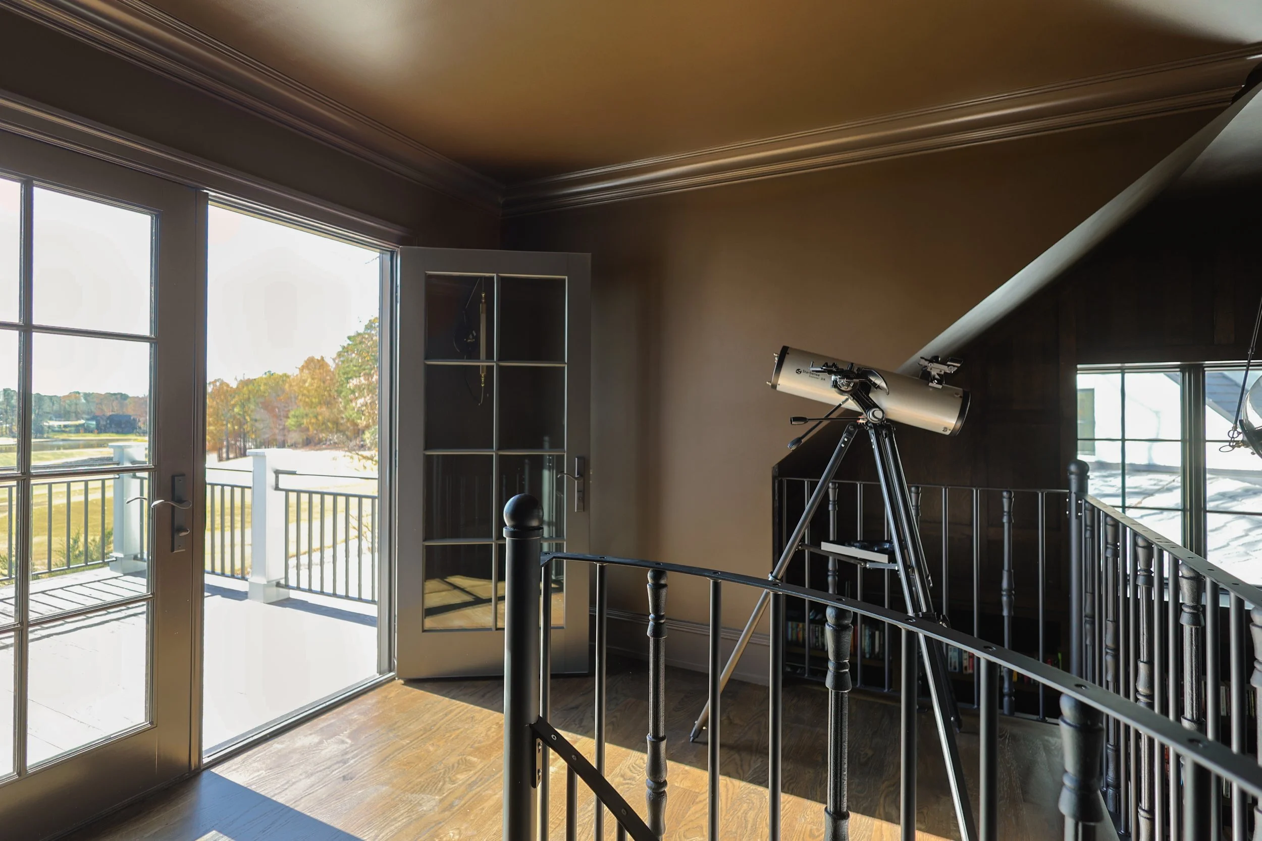 A room with a telescope set up near a large glass door leading to a balcony, overlooking a field and trees outside.