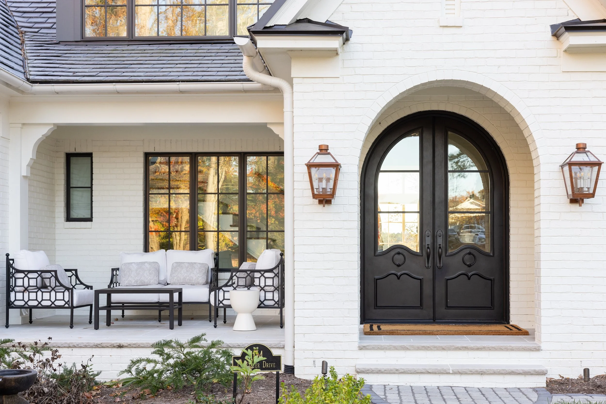 Front porch of a white brick house with black doors, outdoor seating area, and decorative lighting.