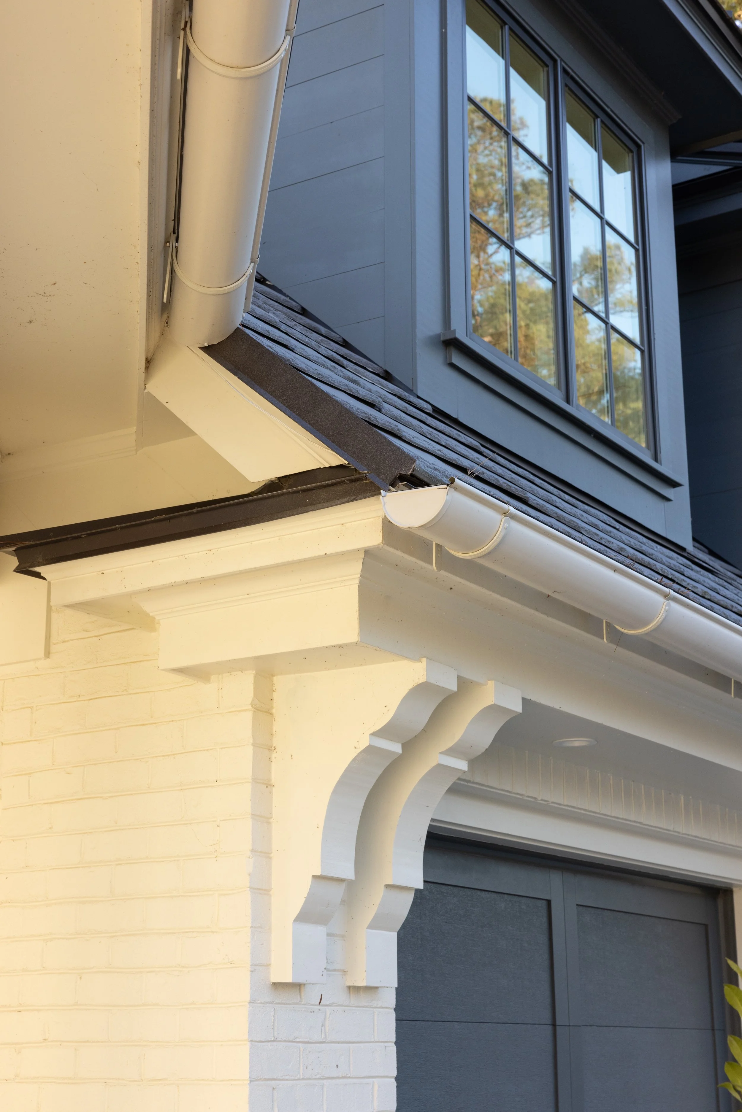 Close-up of the corner of a house showing white trim, grey siding, a window, and gutter system.