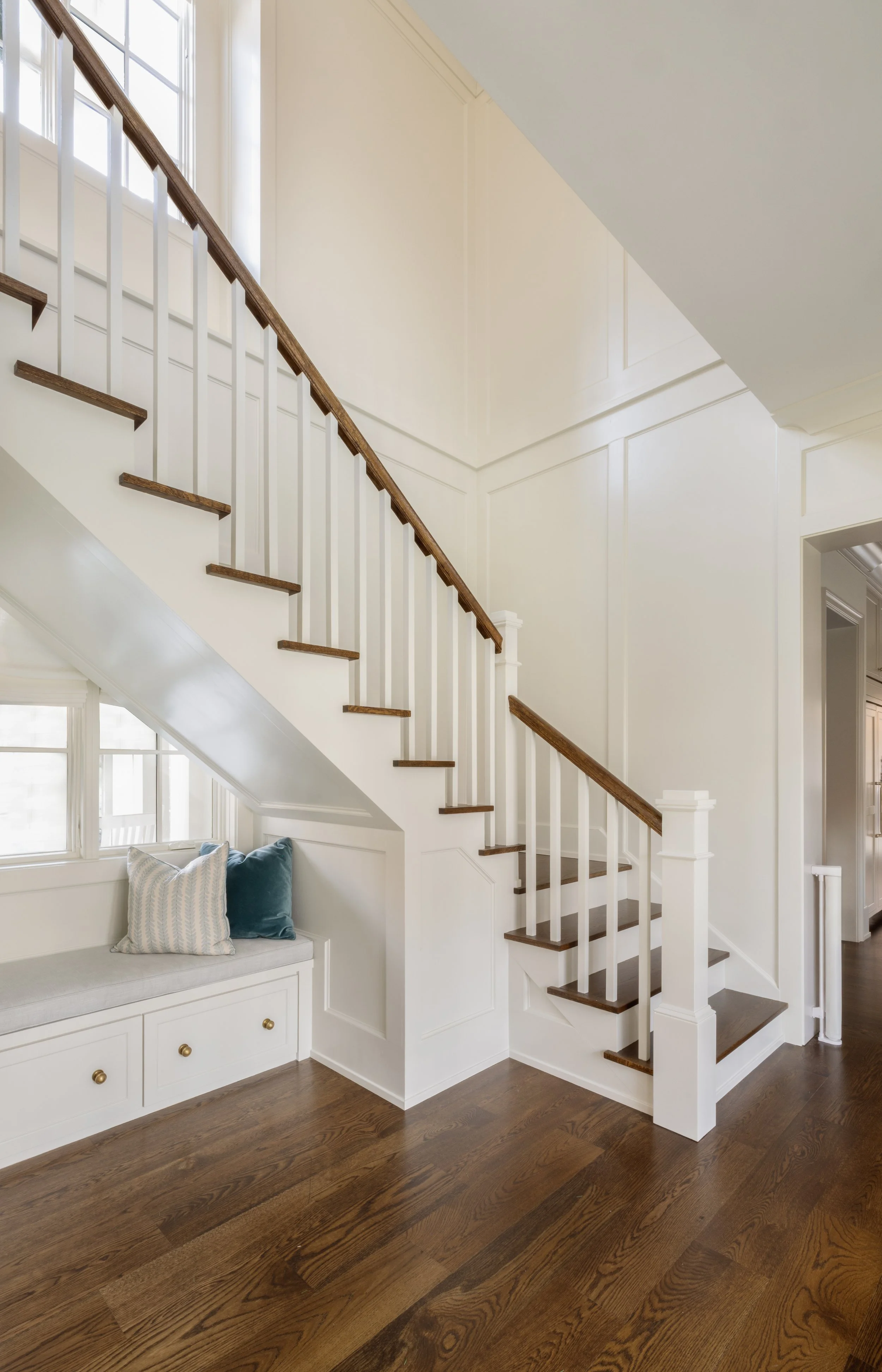 Interior view of a staircase with dark wooden steps and white railing, featuring built-in seating area under the stairs with pillows, dark wooden flooring, and large windows allowing natural light.