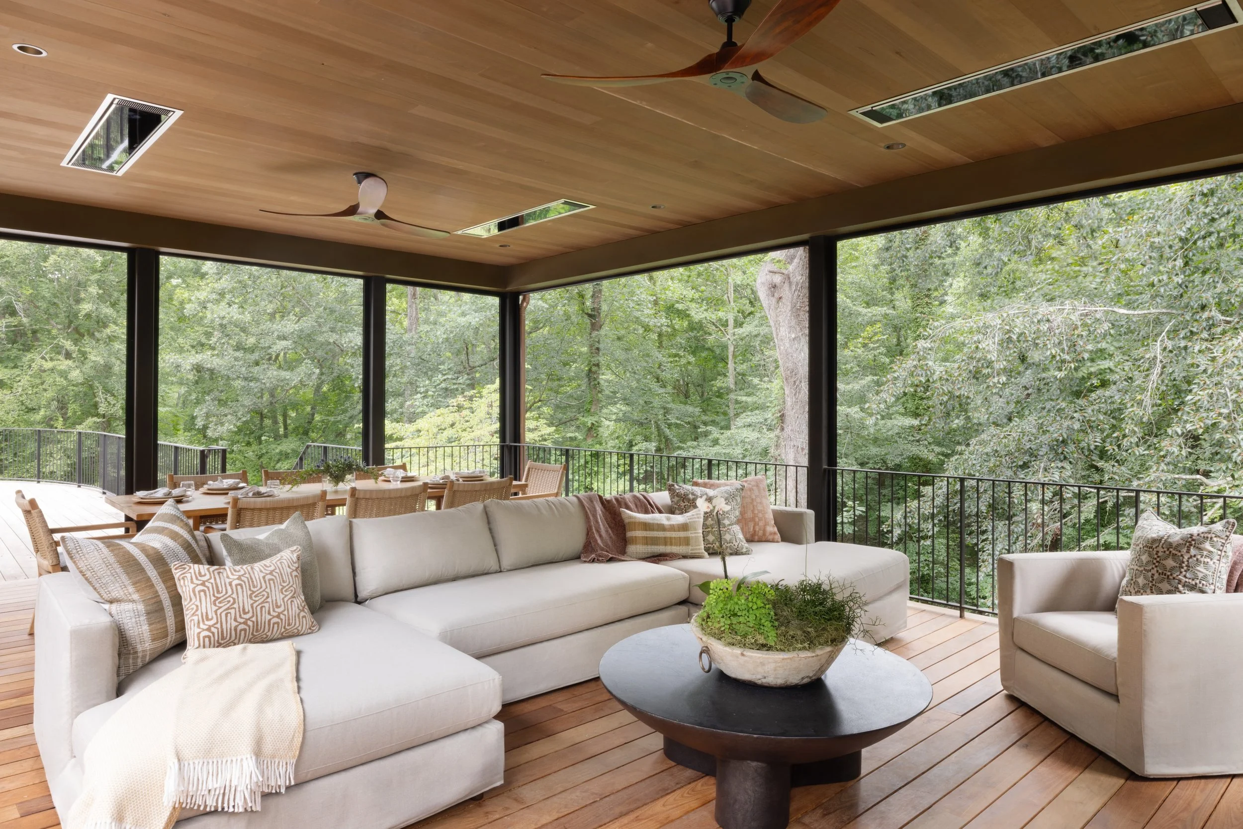 Living room with white sectional sofa, armchair, and wooden coffee table with greenery, overlooking a forest on a screened porch.