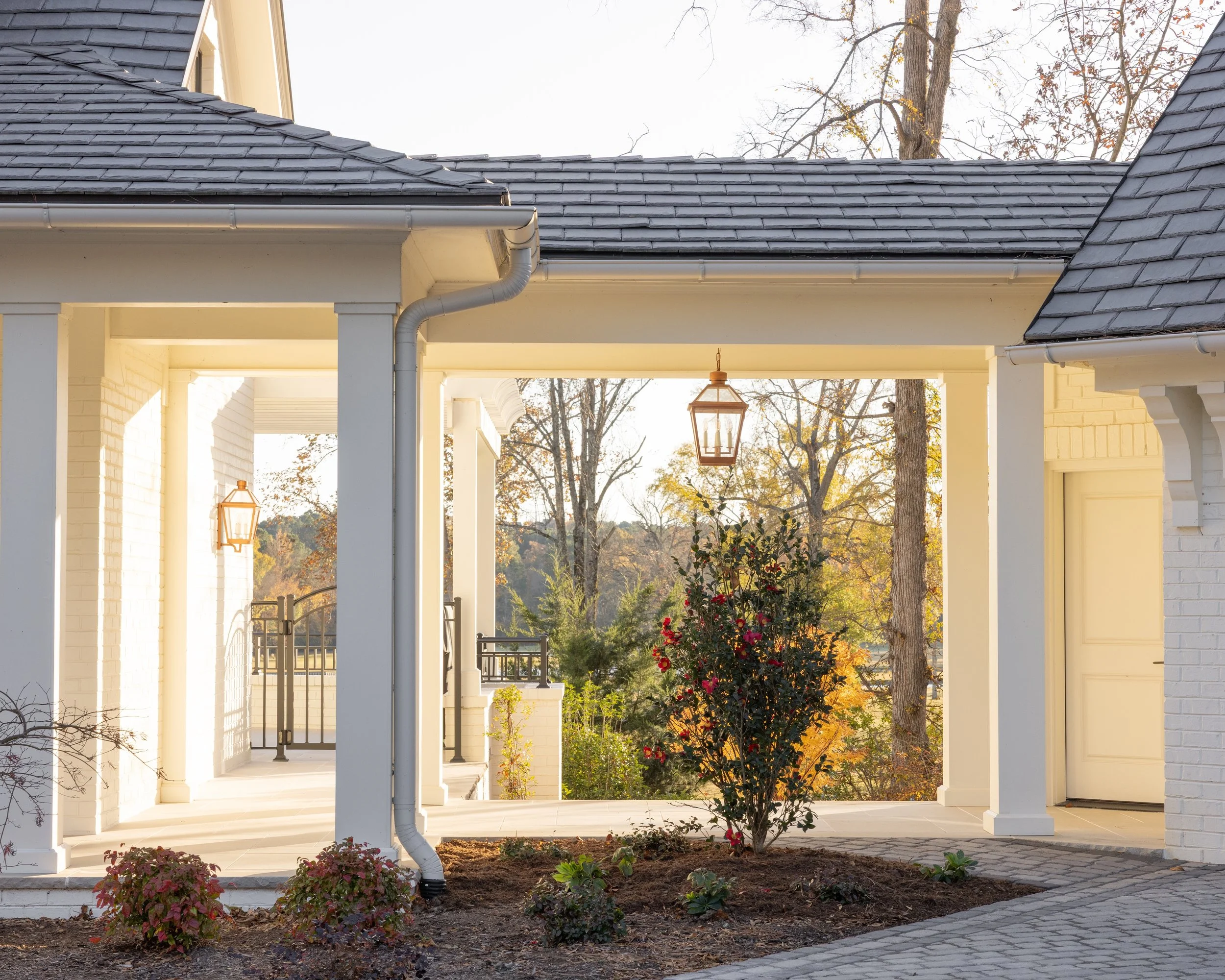 Front porch of a house with white brick walls, black shingle roof, lantern-style hanging lights, and a garden with small plants and bushes.