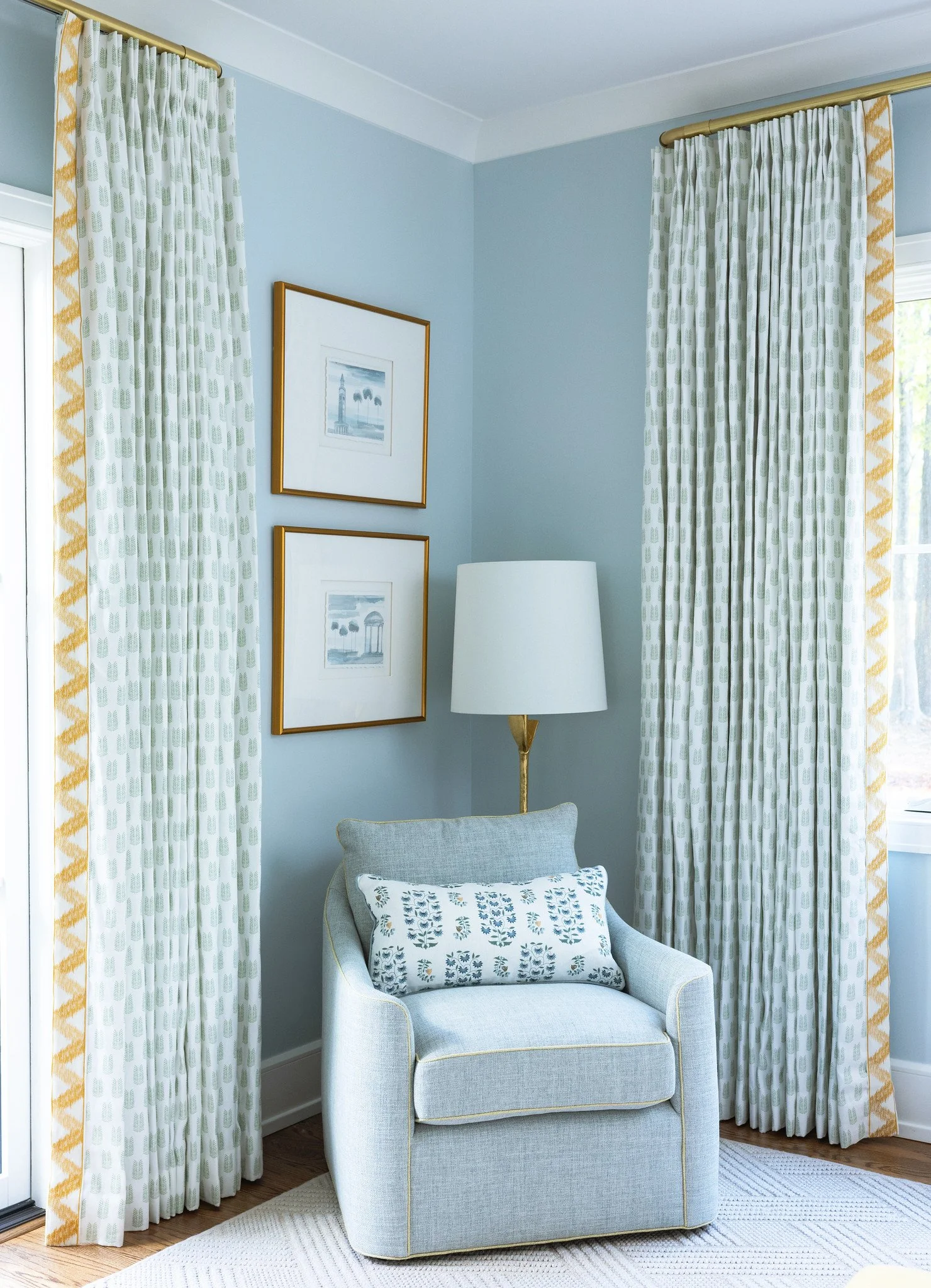 A cozy corner in a living room featuring a light blue armchair with a patterned pillow, a white floor lamp with a gold base, and two framed pictures on the wall. The space is decorated with cream and gold patterned curtains and a light-colored rug.