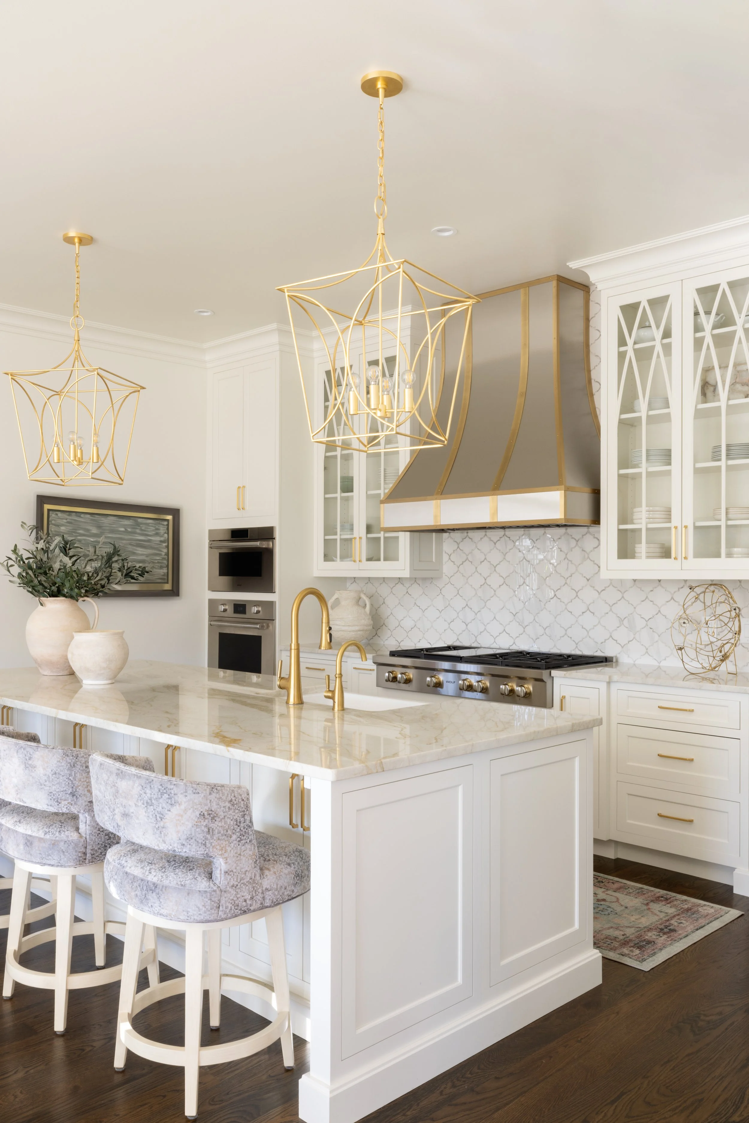 White modern kitchen with gold accents, two gold chandeliers, a marble island with gold fixtures, and glass-front cabinets.