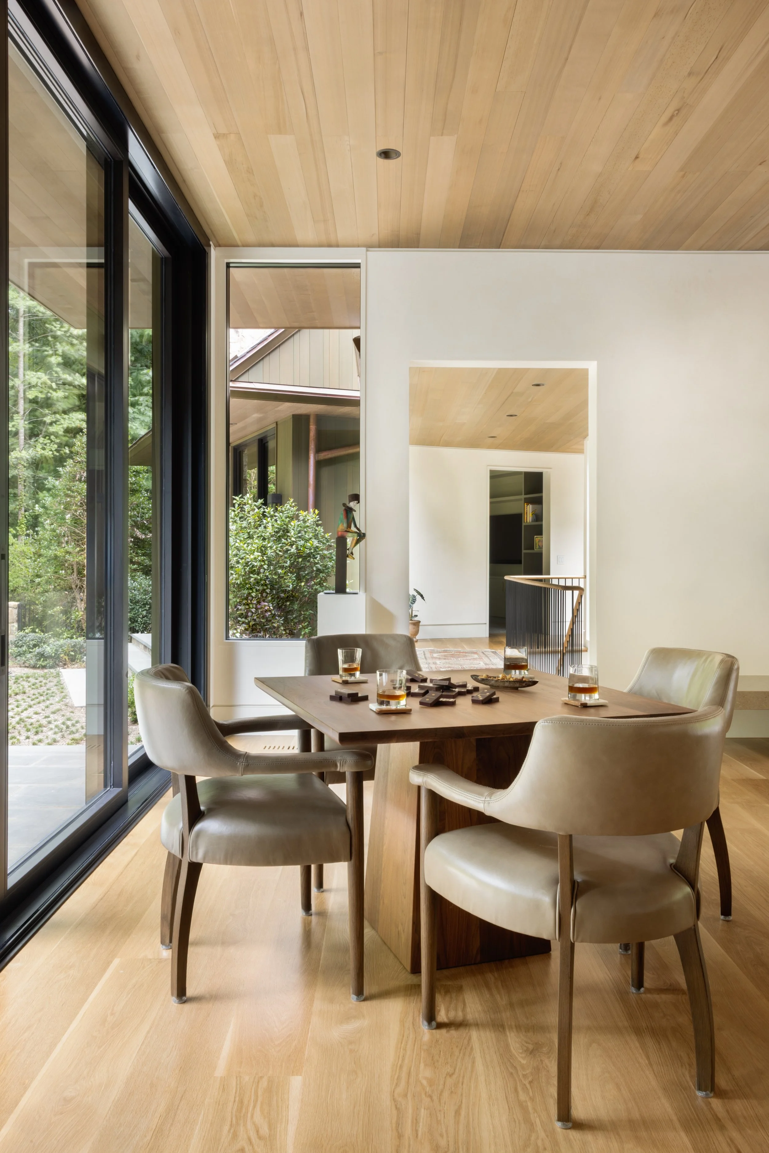 Dining room with a wooden table, four beige upholstered chairs, a glass of drink on each chair, large sliding glass doors, a view of a garden outside, and a staircase through an open doorway inside.