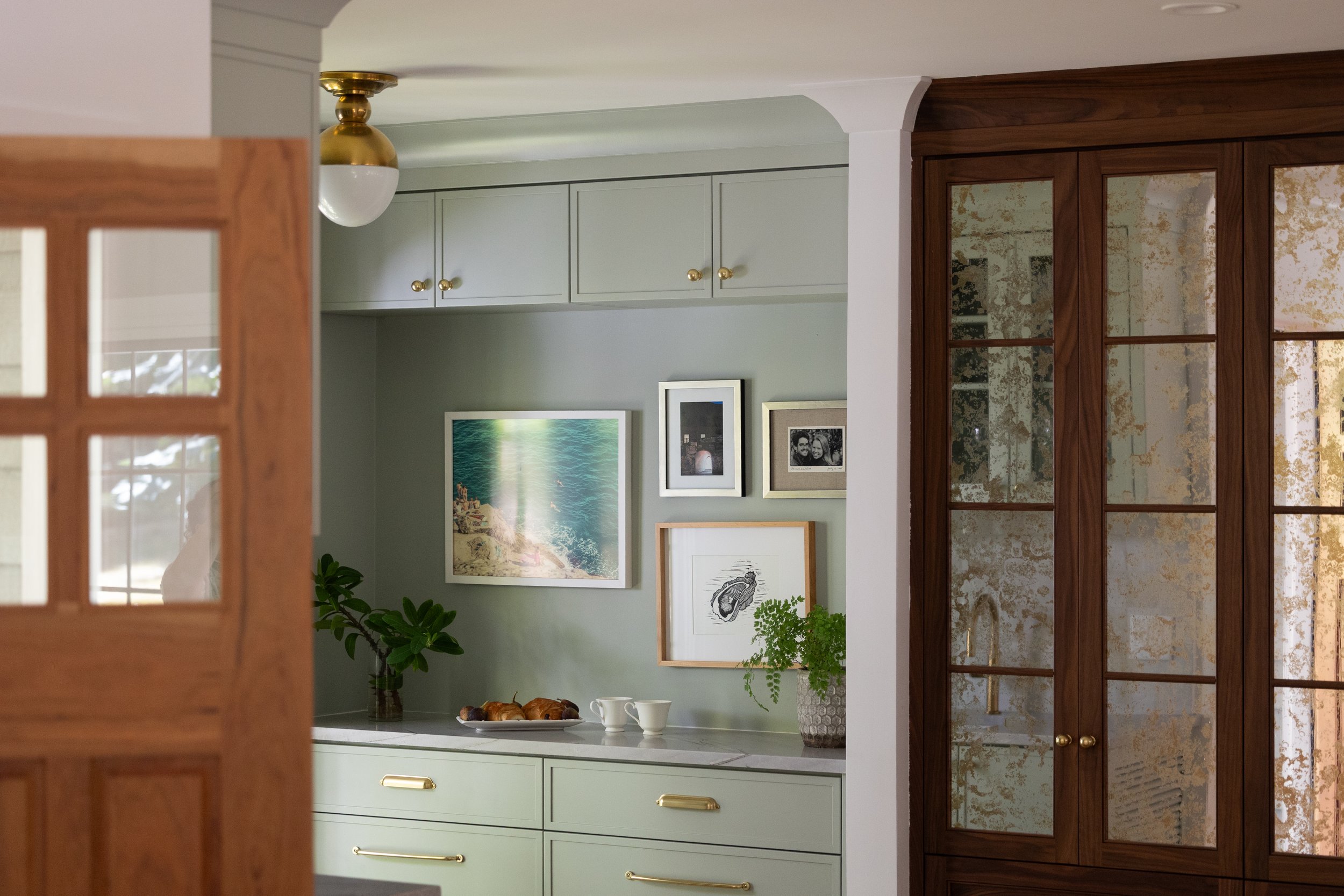 View of a kitchen with mint green cabinets, a wooden cabinet with glass doors, plants, framed artworks, and a tray with croissants on the countertop.