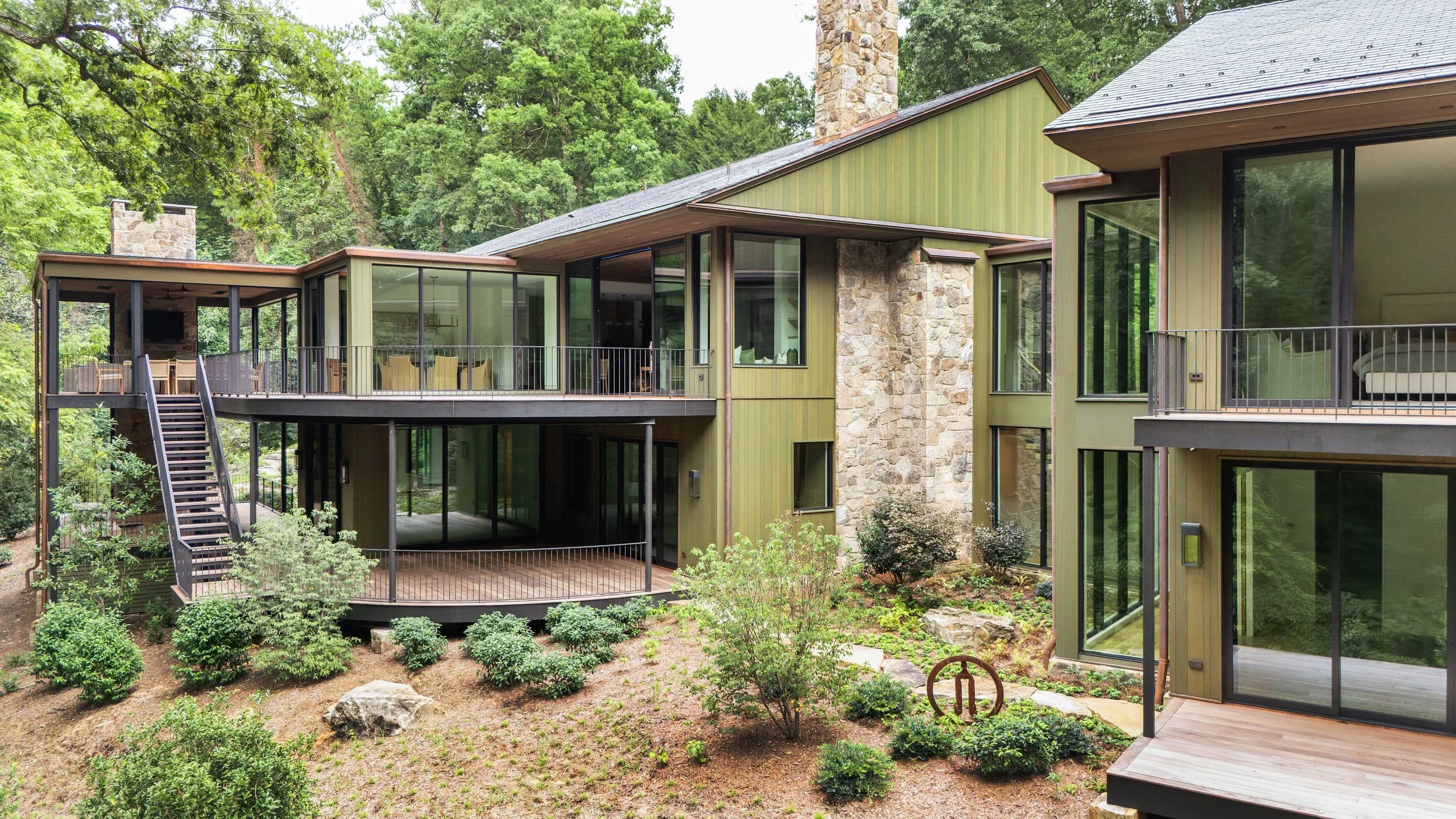 Modern house with large glass windows and multiple balconies, surrounded by green trees and landscaped garden with small shrubs and rocks.
