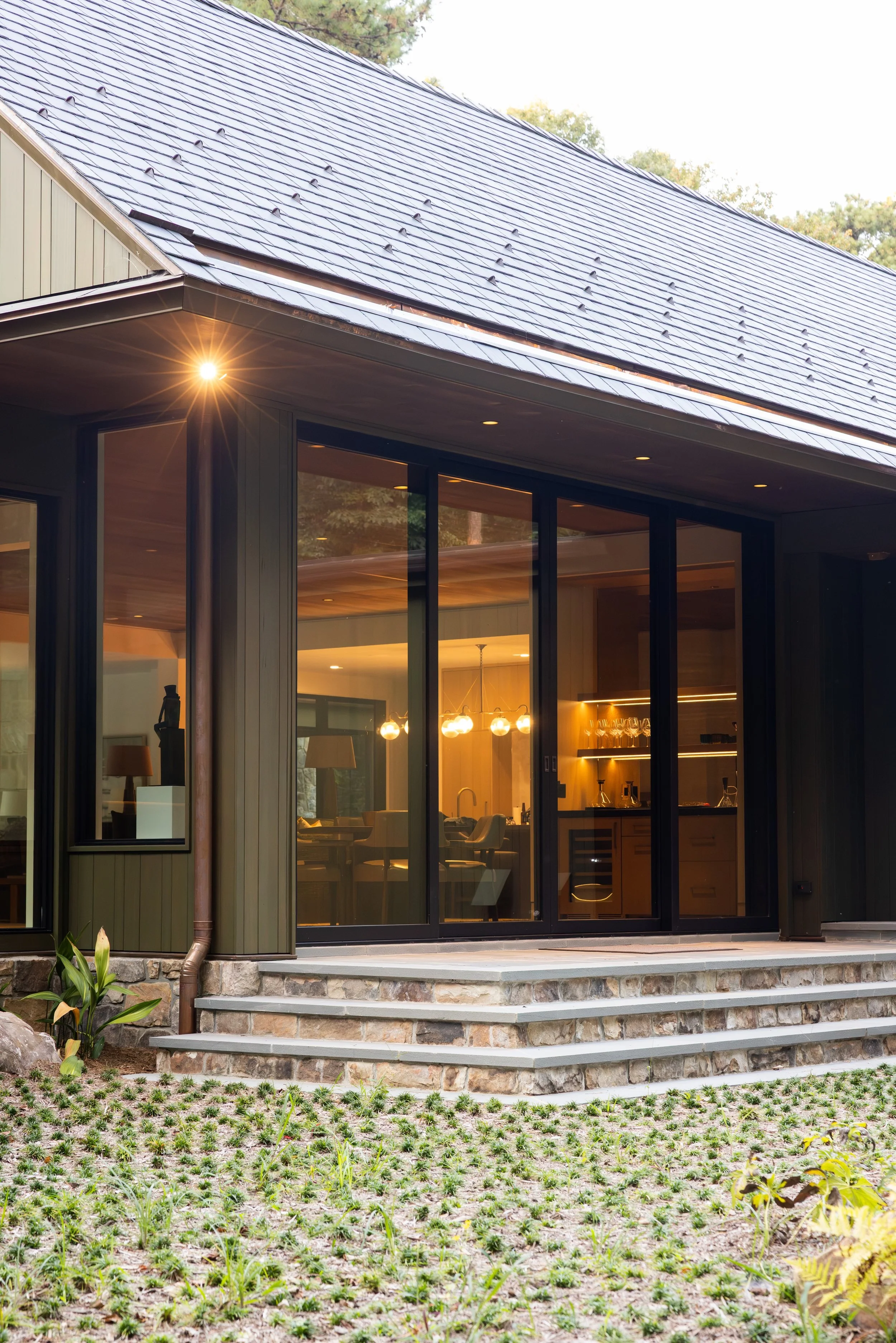 Modern house with large glass sliding doors, interior visible with warm lighting, stone steps leading to the entrance, and a landscaped yard with small plants and flowers.