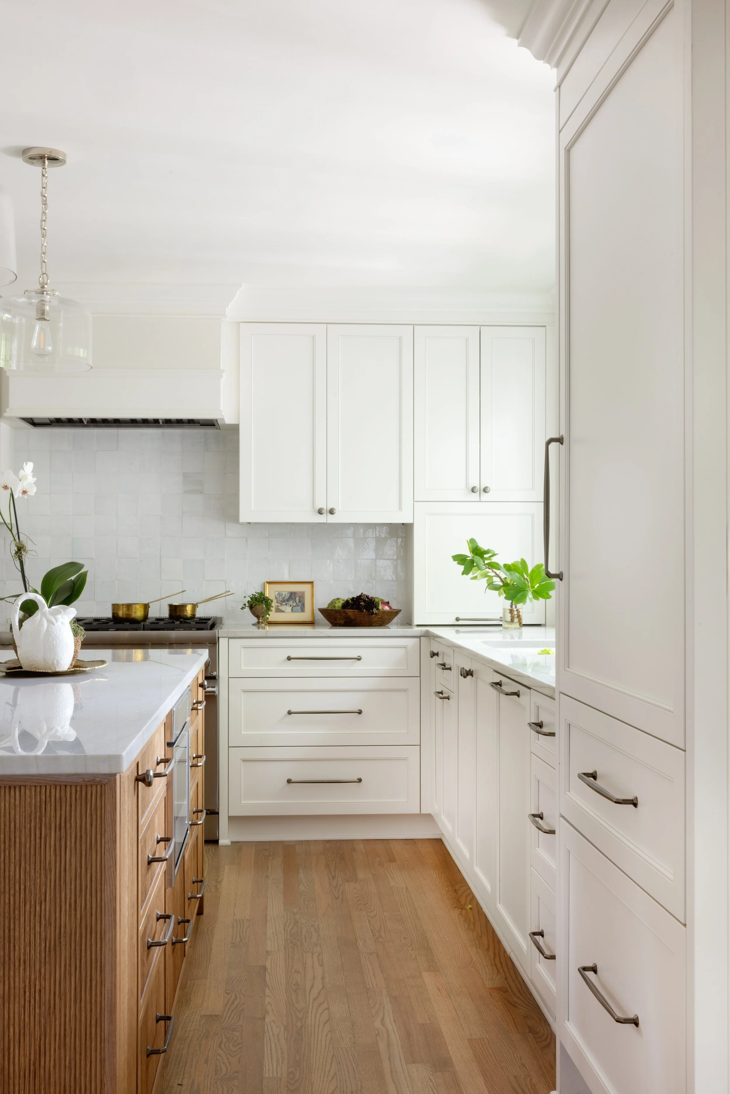 A spacious kitchen with white cabinets, a marble countertop, wooden drawers, and a hardwood floor. There are decorative plants, a framed photo, and a pottery pitcher on the counter.