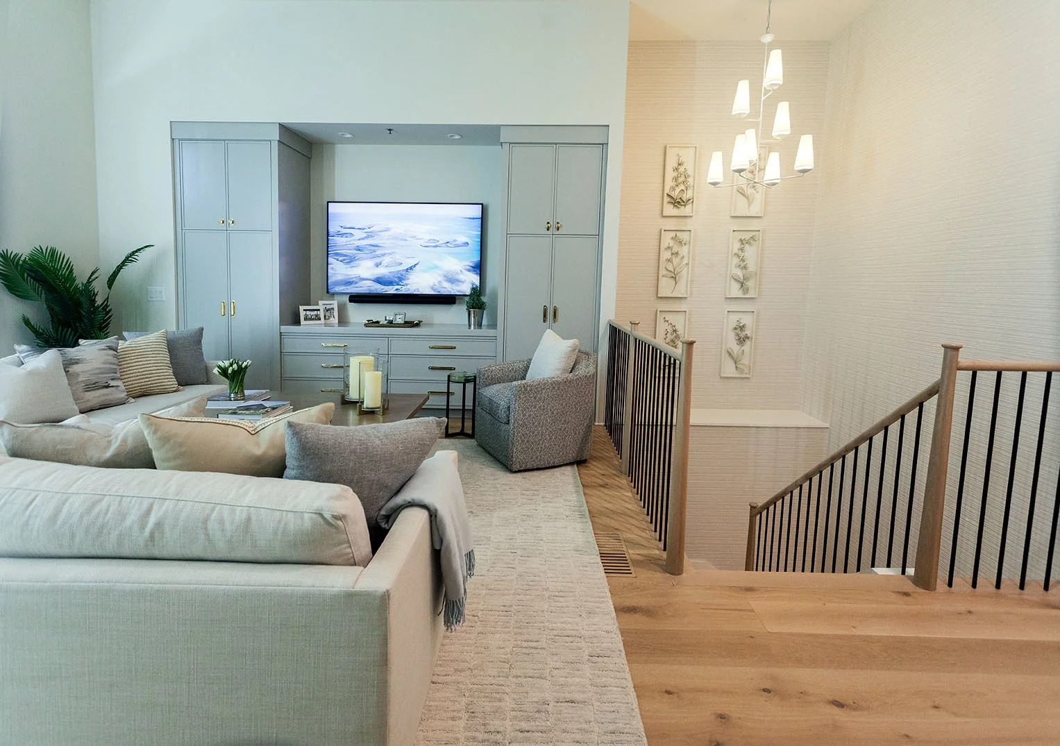 Living room with beige sofa, throw pillows, small armchair, gray built-in cabinets with TV, decorative candles, and wall art, with hardwood flooring and a staircase with black railing and wooden handrail.