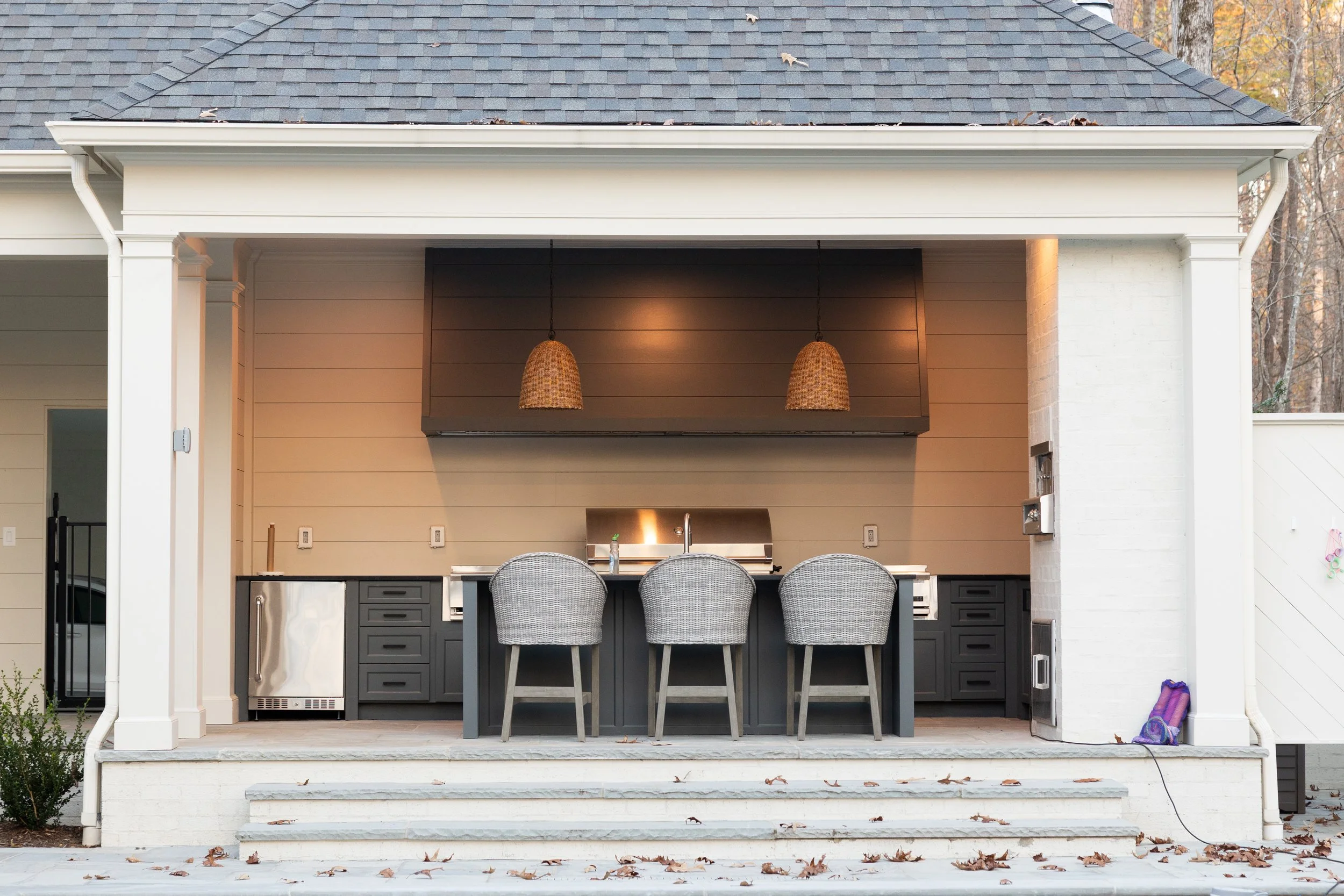 Outdoor kitchen with a countertop, three chairs, and two hanging pendant lights under a covered patio.