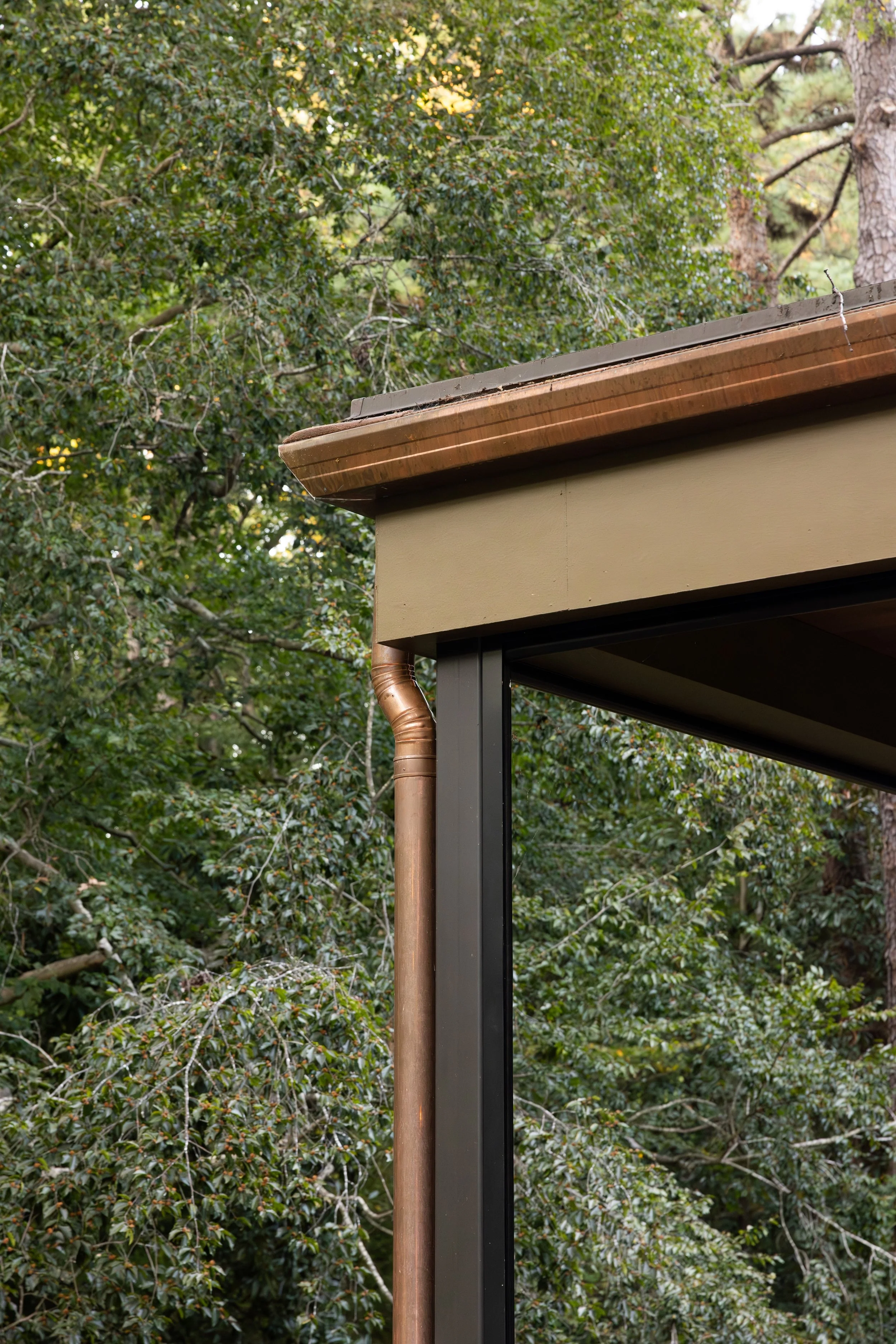 Close-up of the corner of a house with a brown gutter, beige fascia, and black metal support beams, with green leafy trees in the background.