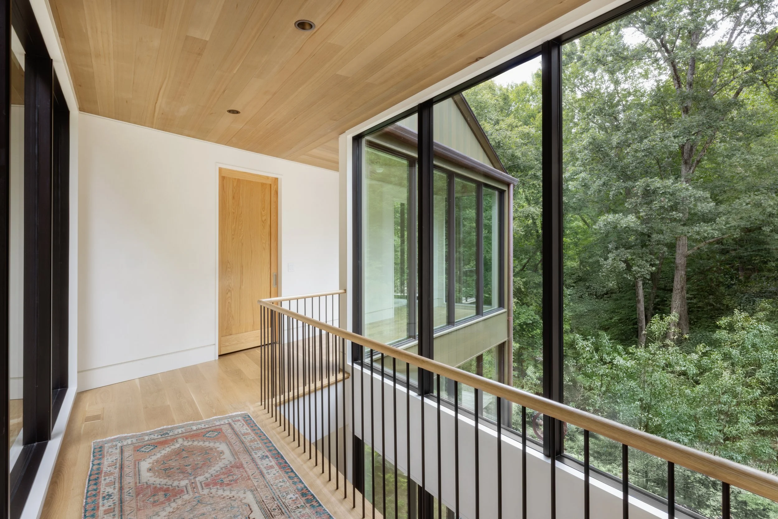 Interior view of a modern house hallway with wooden floors, a colorful rug, a white wall, a wooden door, and a large floor-to-ceiling glass window overlooking trees outside.