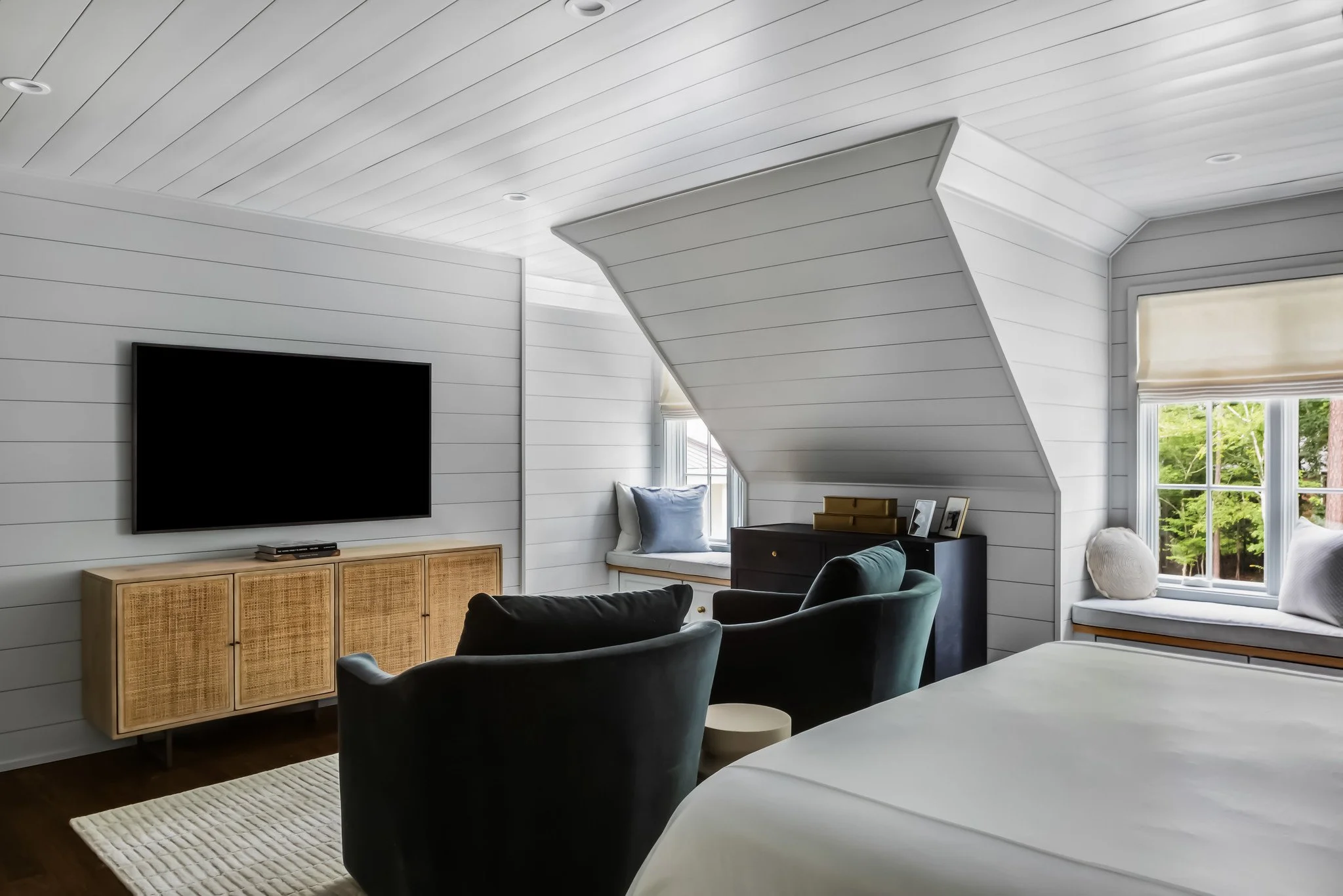 A bedroom with white wooden walls and ceiling, a large flat-screen TV on a wooden cabinet, two black armchairs, and a bed with white bedding near windows with green trees outside.