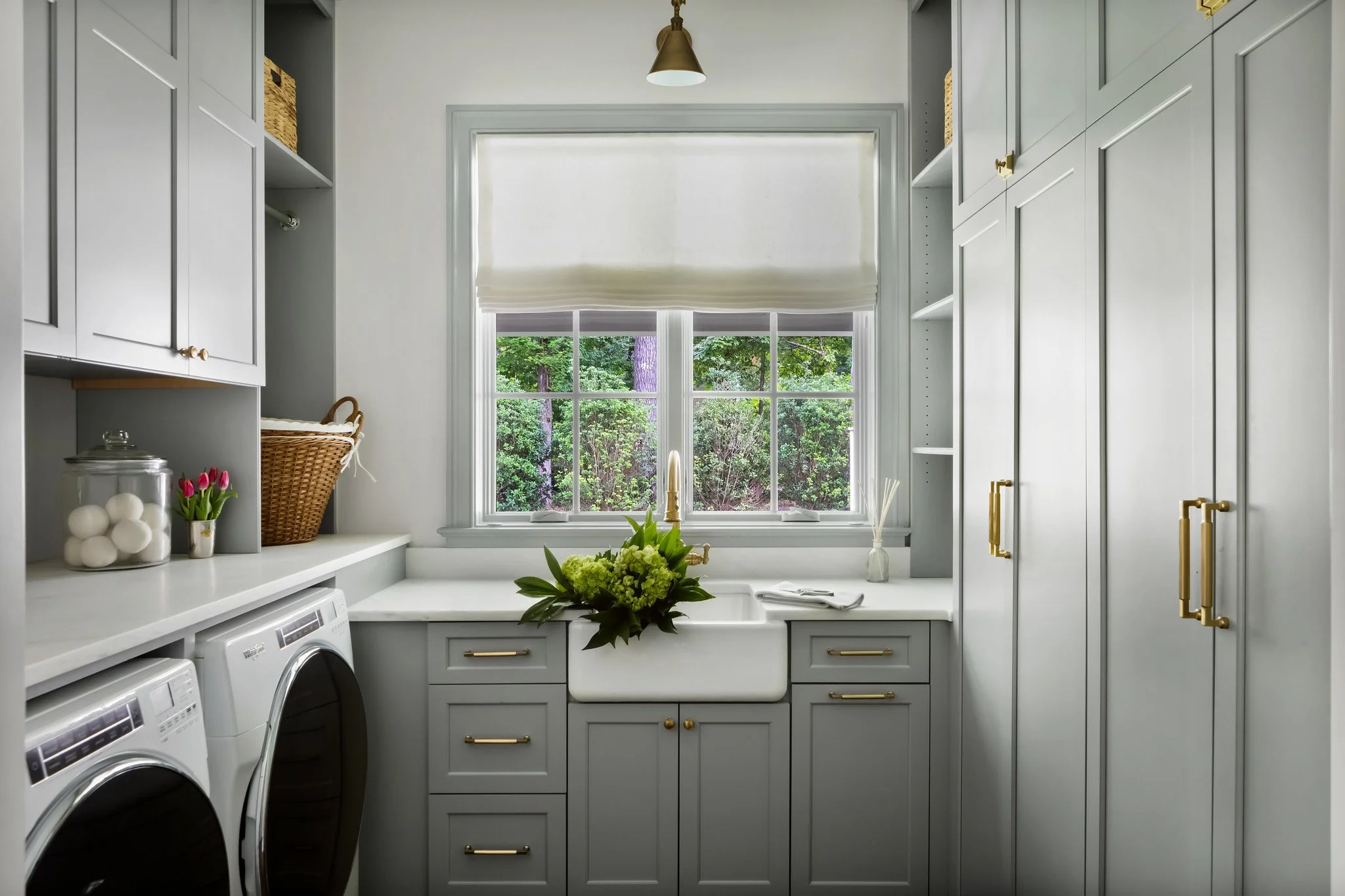 A laundry room with grey cabinets, a white farmhouse sink with a gold faucet under a window with a view of green trees, and a countertop with a flower arrangement. There are washing machines on the left and open shelves on the right.