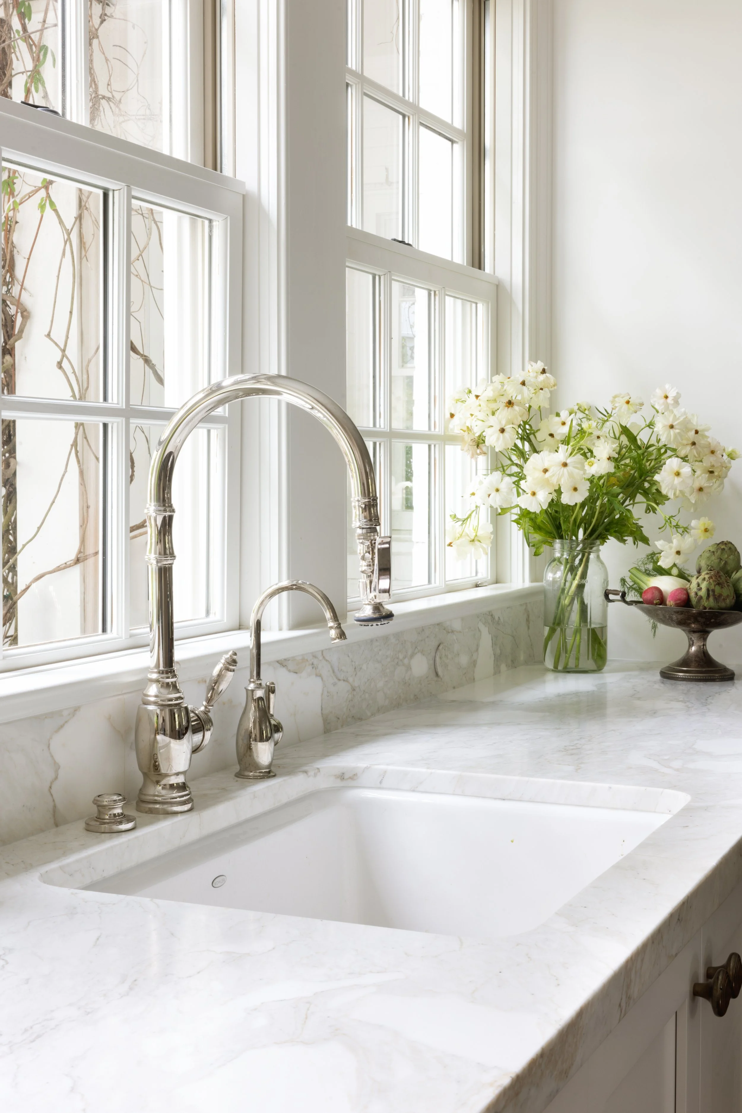 White marble kitchen sink with chrome faucet, large window with multiple panes, vase of white flowers, and a bowl of radishes and artichokes on the countertop.