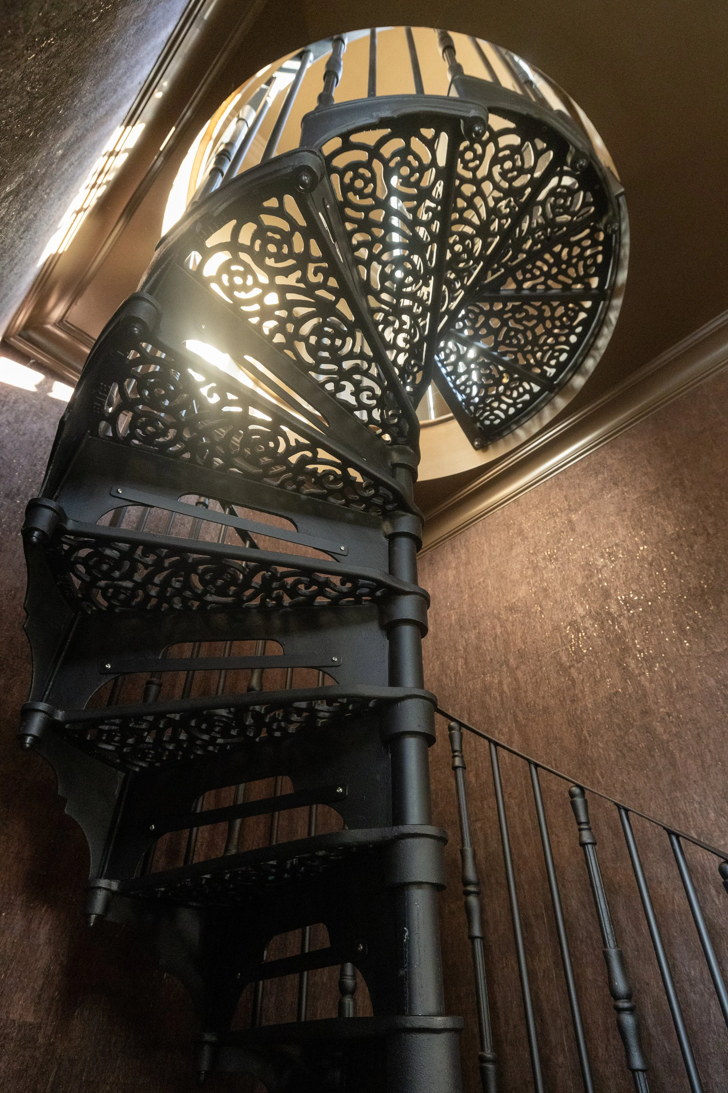 A spiral staircase with ornate black metalwork and a wooden handrail, viewed from below, with sunlight shining through the open center.