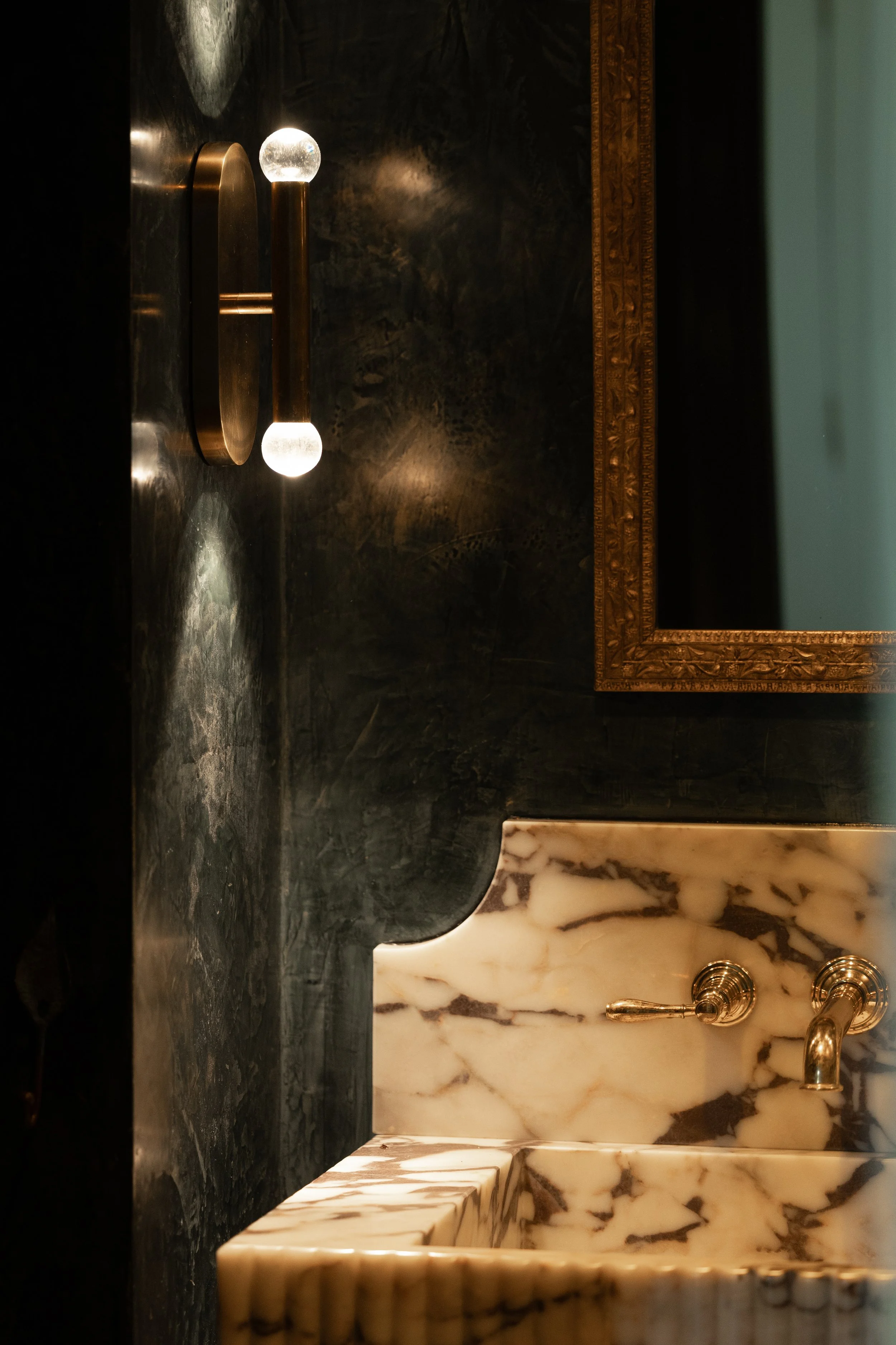 Close-up of a bathroom sink with a marble basin, a brass faucet, a dark marble wall, a mirror with a gold frame, and wall-mounted light fixtures.