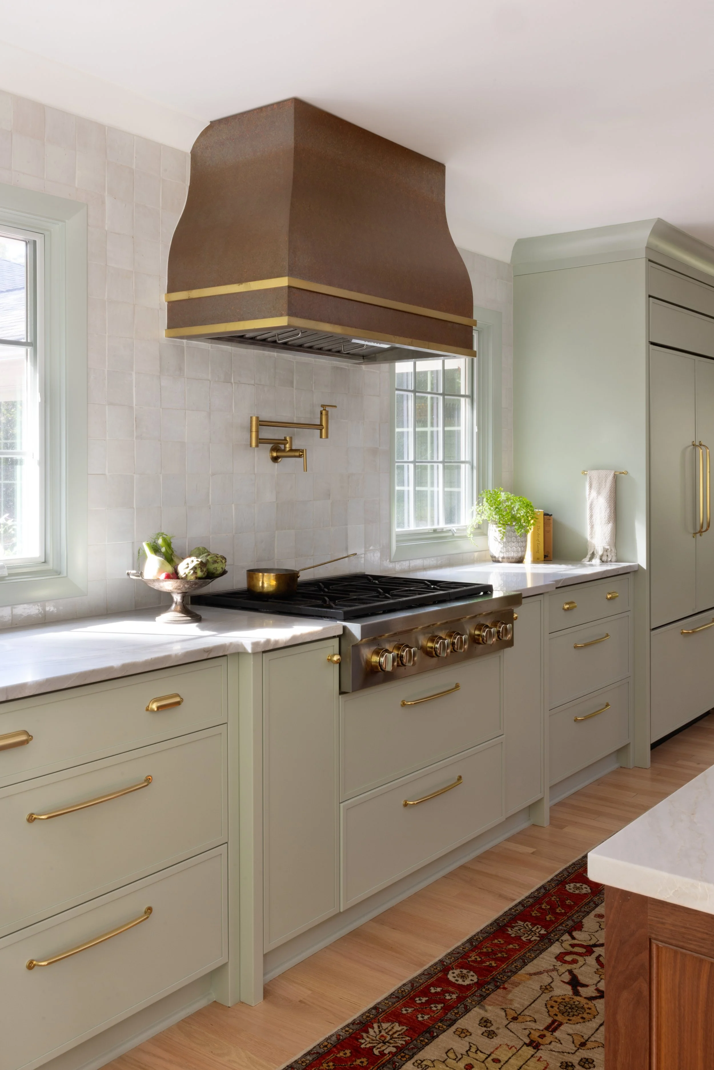 Kitchen with light green cabinets, a gas stove, brass handles, a copper range hood, windows, and a potted plant.