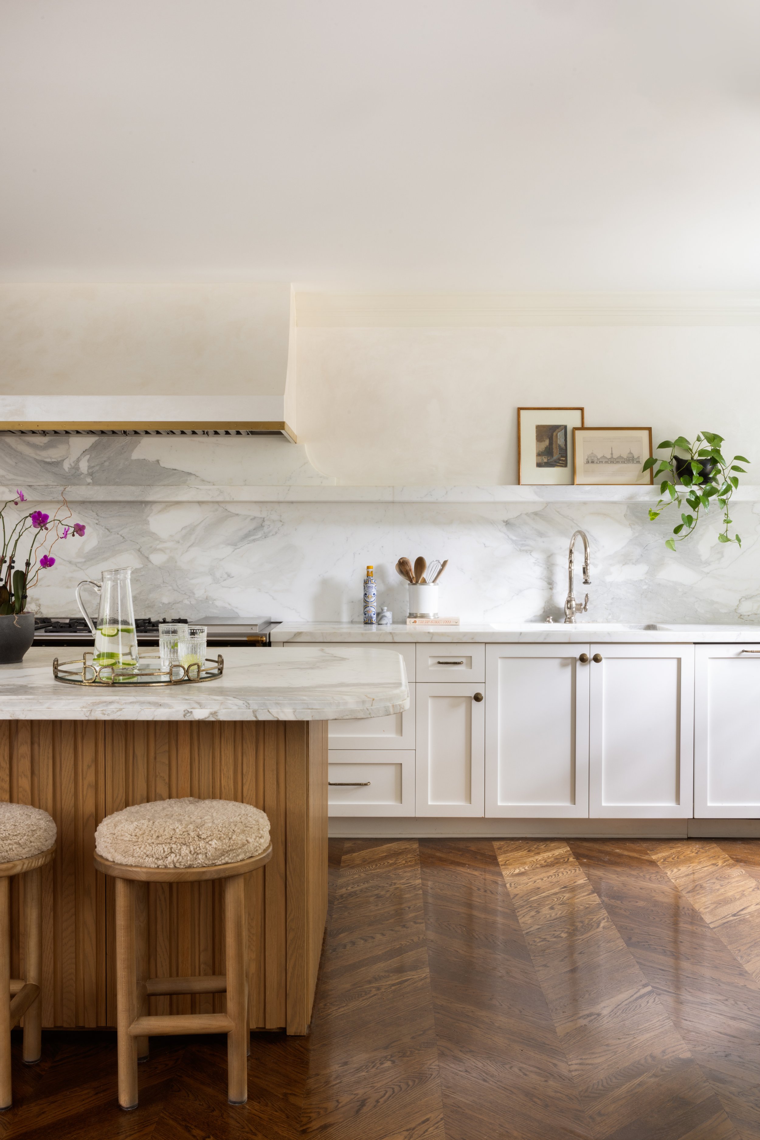 A modern kitchen with white cabinets, a marble backsplash, and a wooden floor. There are framed pictures and a green plant on the shelf, a faucet over the sink, and a kitchen island with two wooden stools with plush seats.