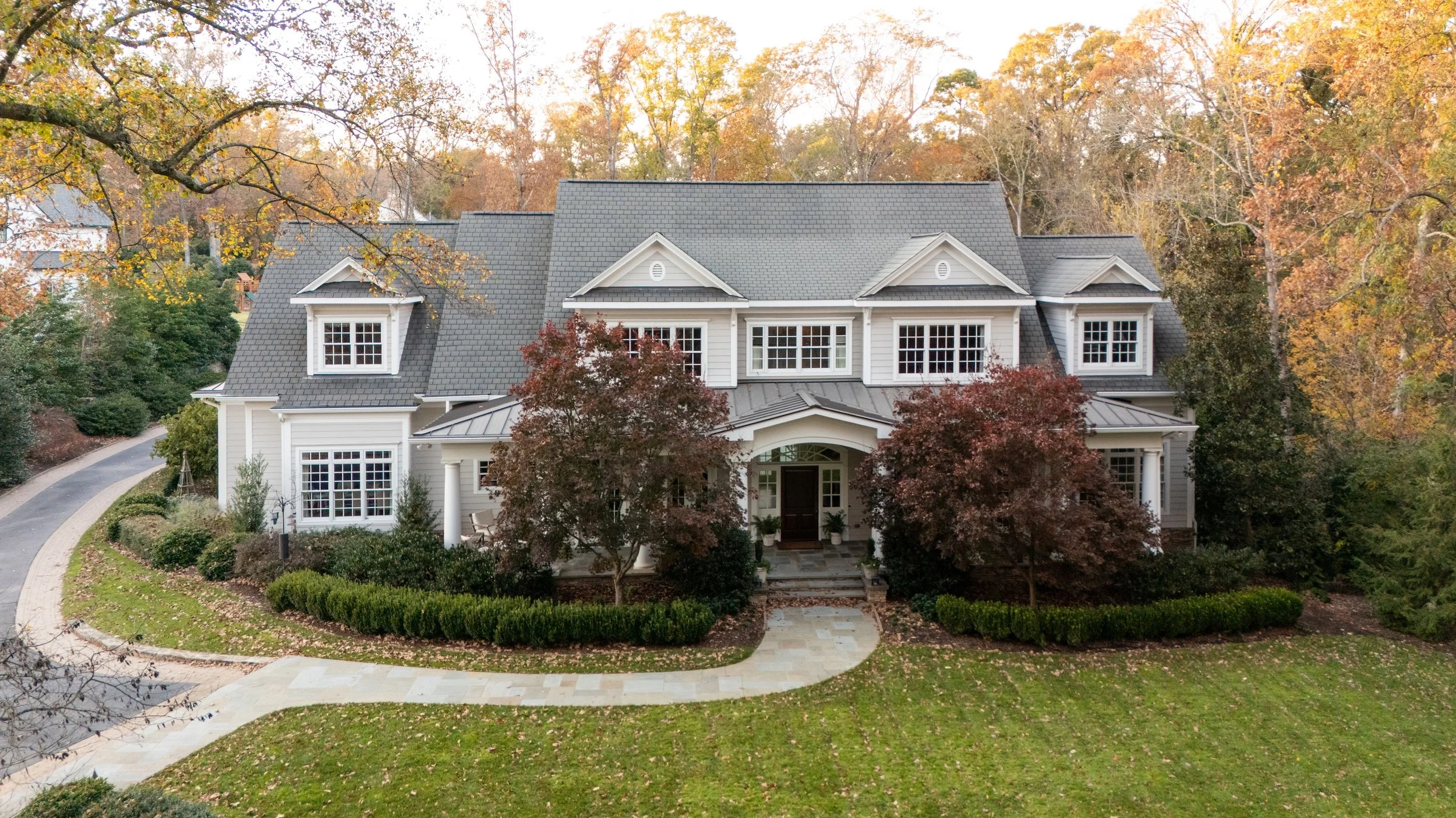 A large, elegant two-story house with white exterior and gray roof, surrounded by trees with autumn foliage and a well-maintained lawn with a curved walkway leading to the front entrance.