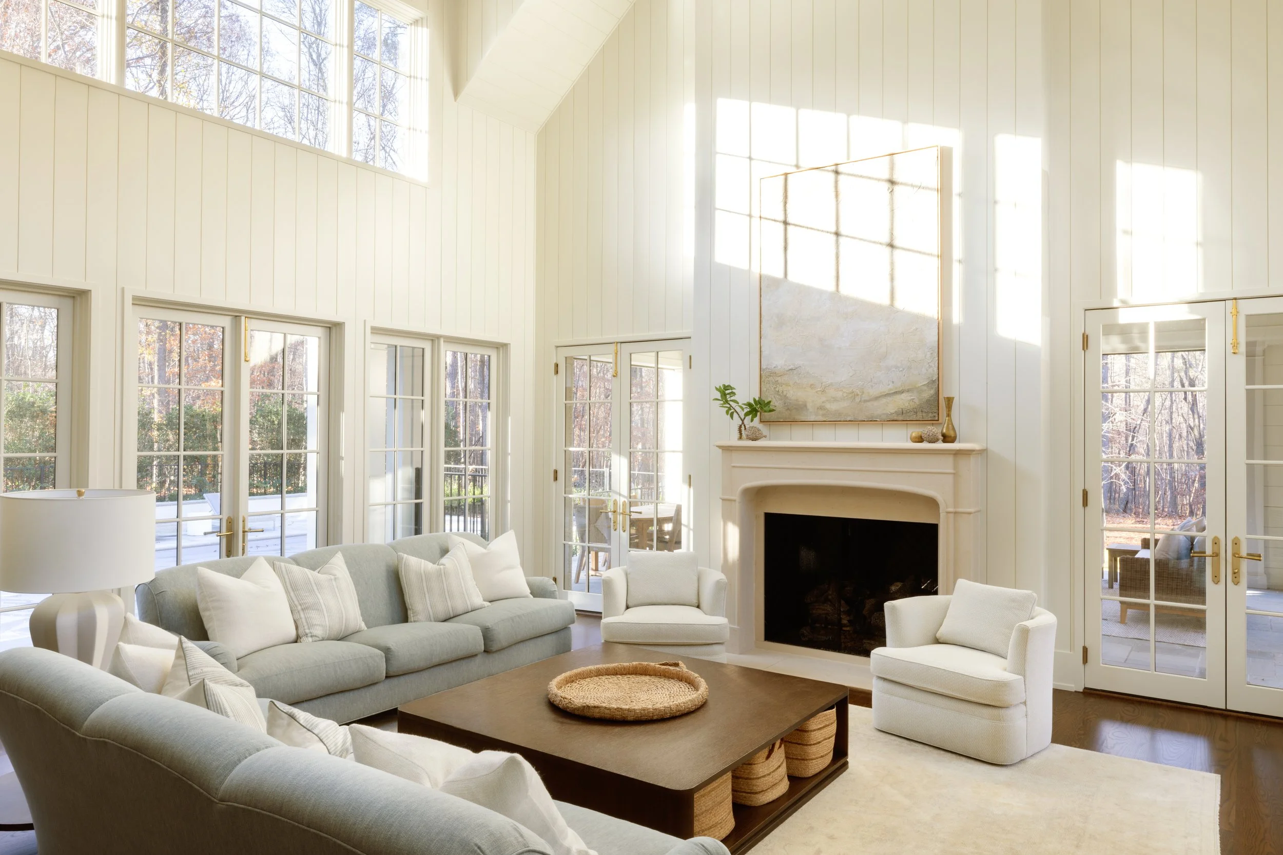 Bright living room with white paneled walls, large windows, a fireplace, and comfortable seating including a sofa and armchairs, with a wooden coffee table and decor on the mantel.