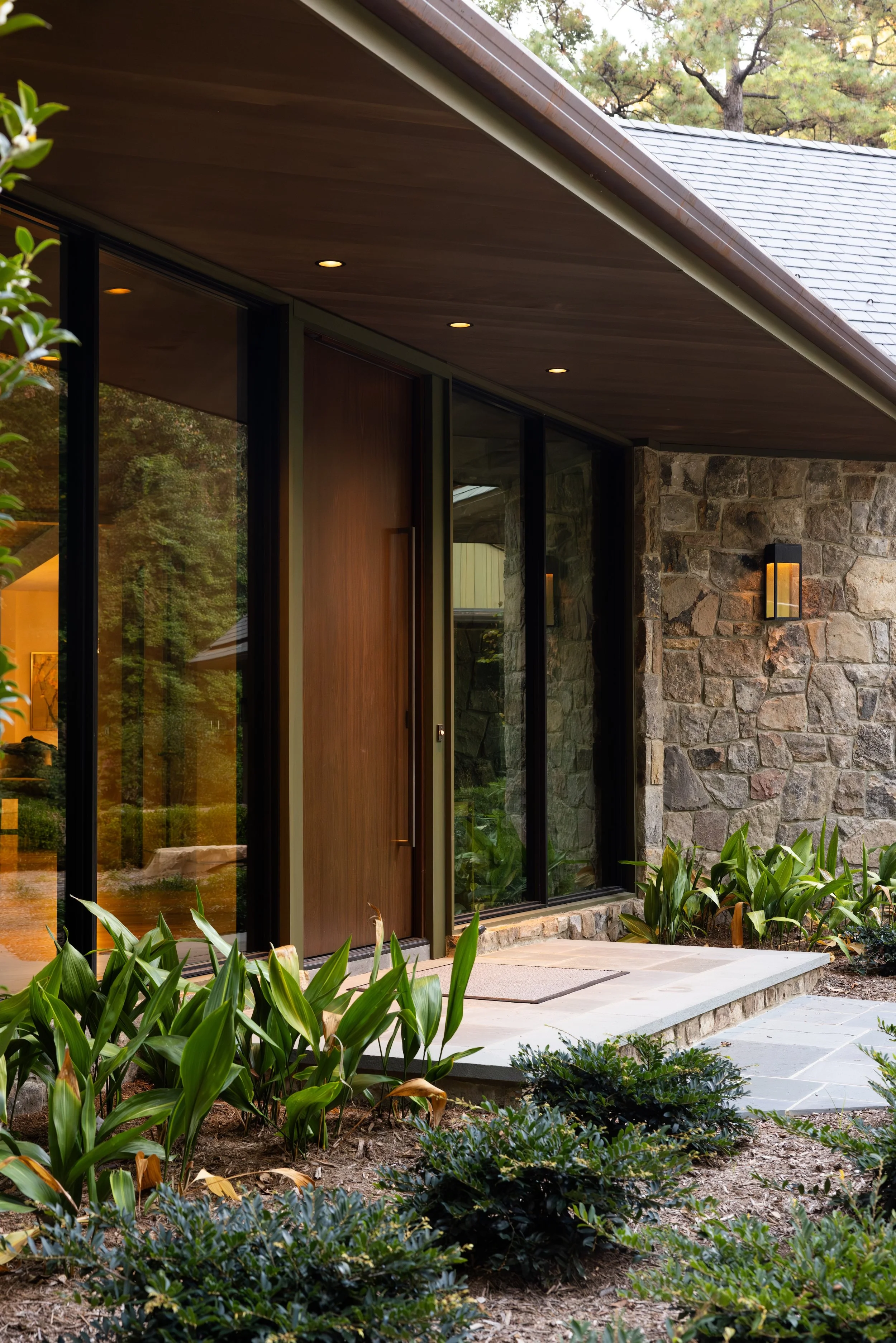 Modern house entrance with large glass windows, a wooden door, stone wall, outdoor lighting, and landscaped garden with green plants.