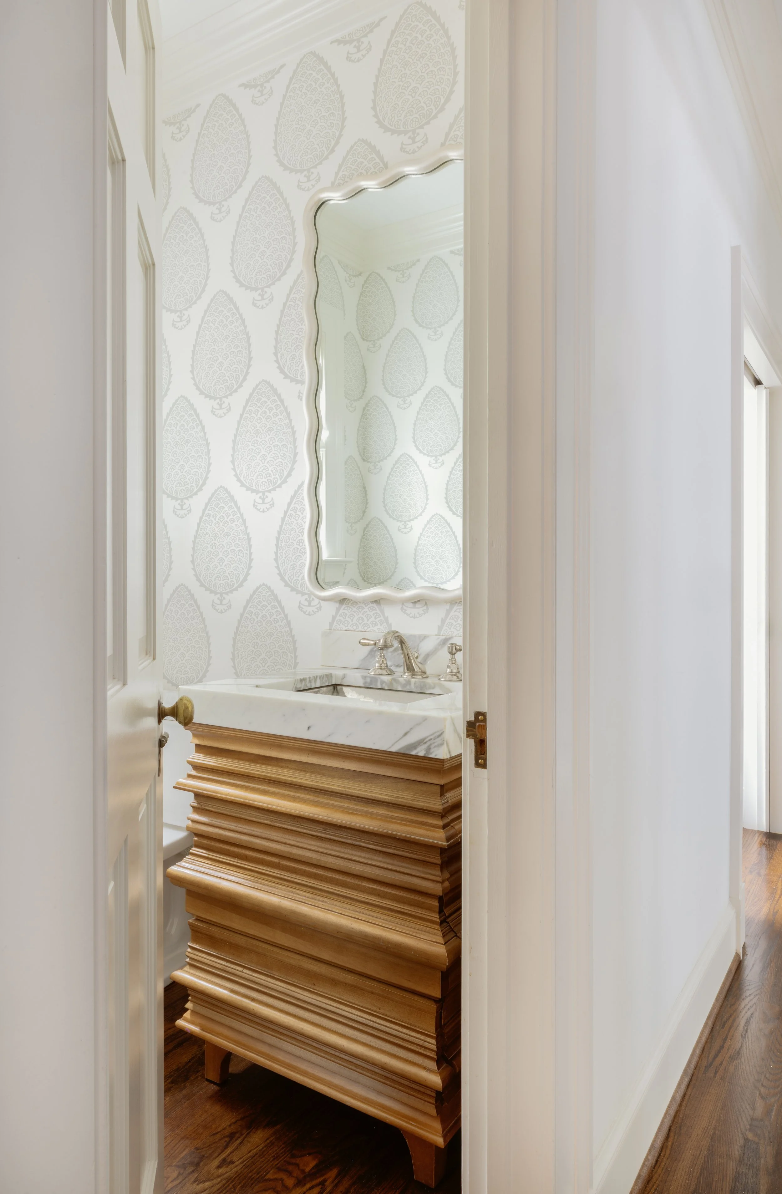 A small bathroom with a marble sink, ornate mirror, patterned wallpaper, and wooden cabinetry, viewed through an open door.