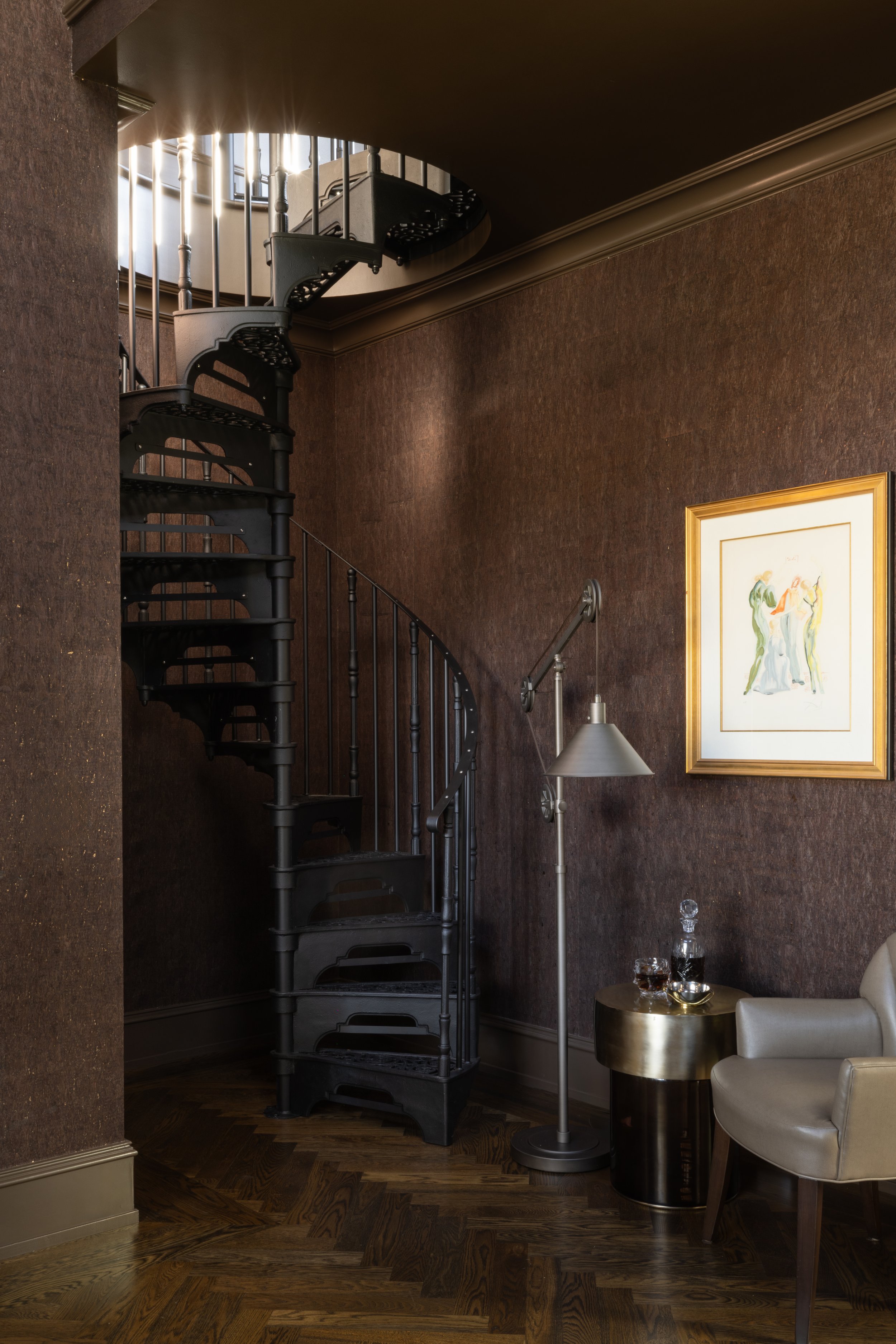 Interior view of a room with a black spiral staircase, a framed abstract artwork on the wall, a beige armchair, a metallic side table with a decanter and glasses, a modern floor lamp, and dark wood flooring.